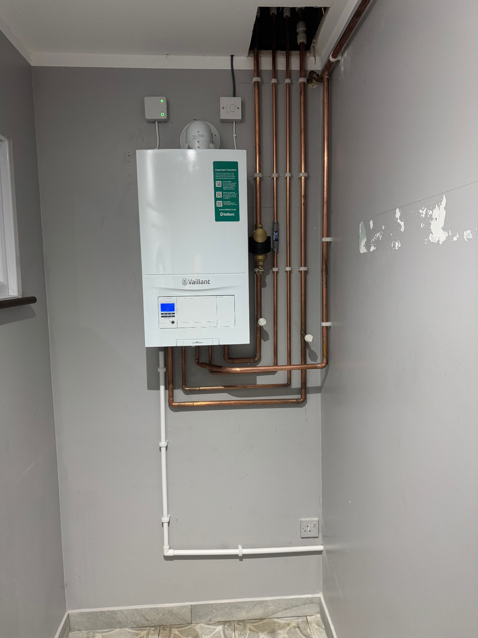 Biały piec gazowy Vaillant zamontowany na szarej ścianie z widocznymi miedzianymi rurami instalacji. Widoczne elementy instalacji elektrycznej i gazowej.