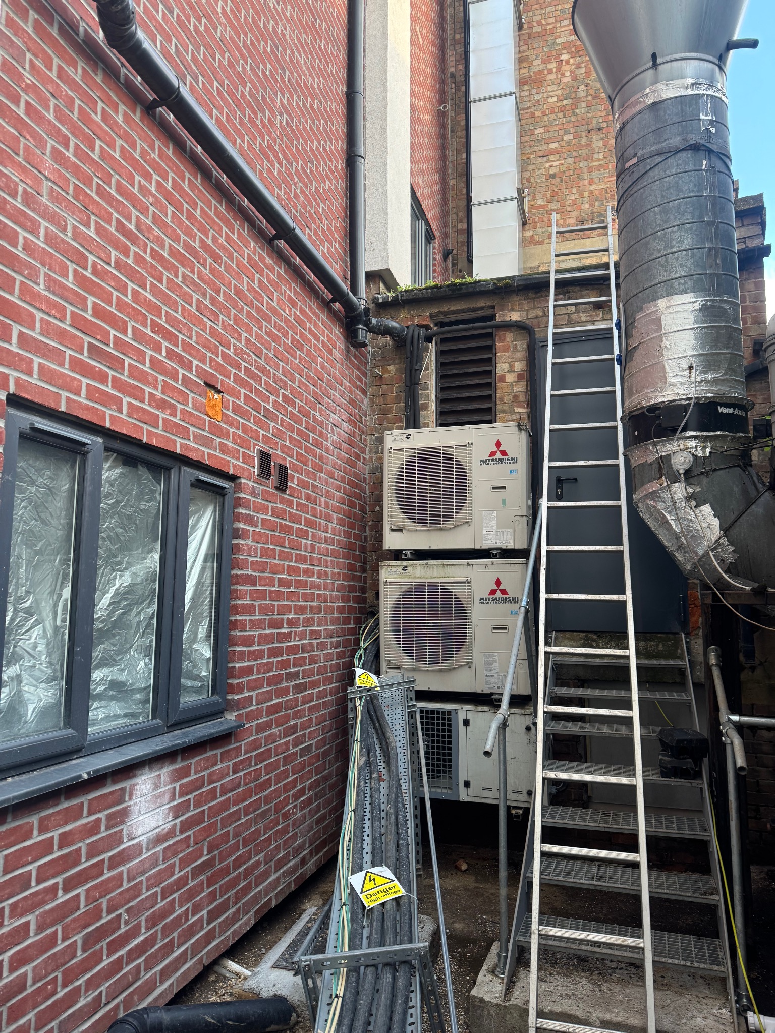 Dwa agregaty klimatyzacyjne Mitsubishi zamontowane na zewnątrz budynku z cegły, obok metalowej drabiny i rur wentylacyjnych. Widoczne kable i ostrzeżenie o wysokim napięciu.