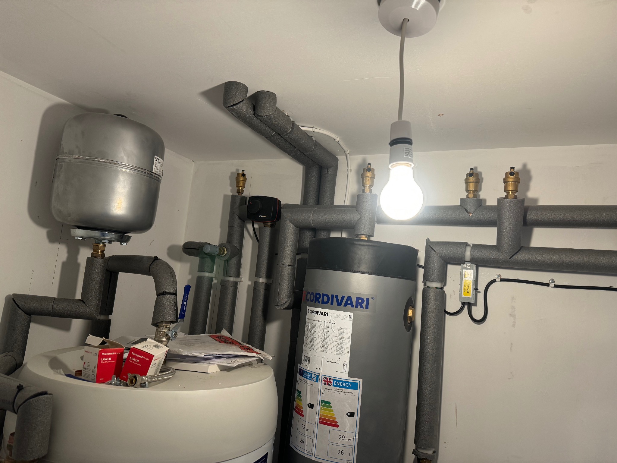 Instalacja CO z zbiornikami Cordivari, rurami w izolacji i elementami montażowymi Honeywell. Widoczne przyłącza i zawory. Oświetlenie żarówką.