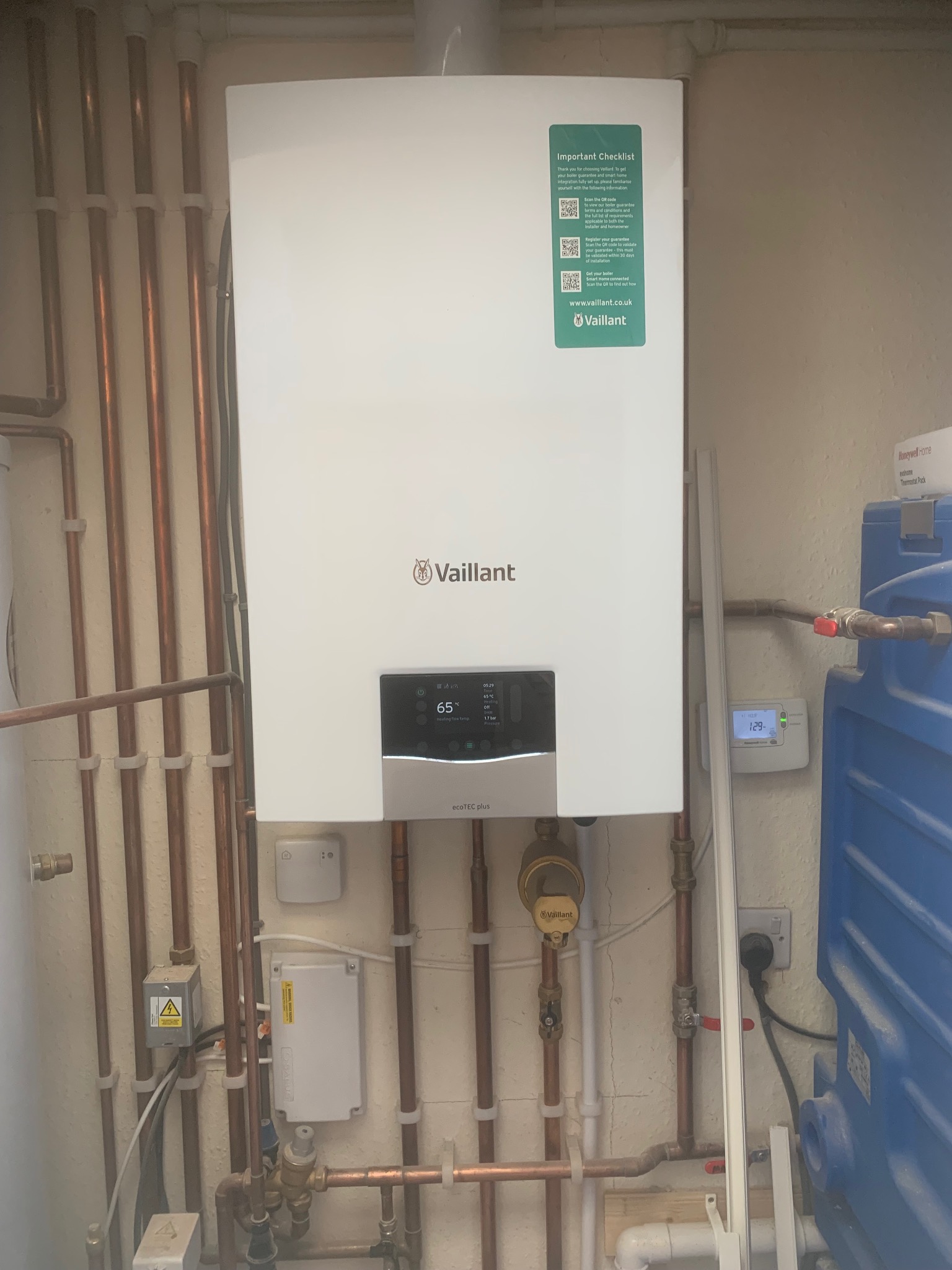 Biały piec gazowy Vaillant zamontowany na ścianie z widocznymi miedzianymi rurami instalacji. Na piecu naklejka z kodami QR i adresem strony internetowej.