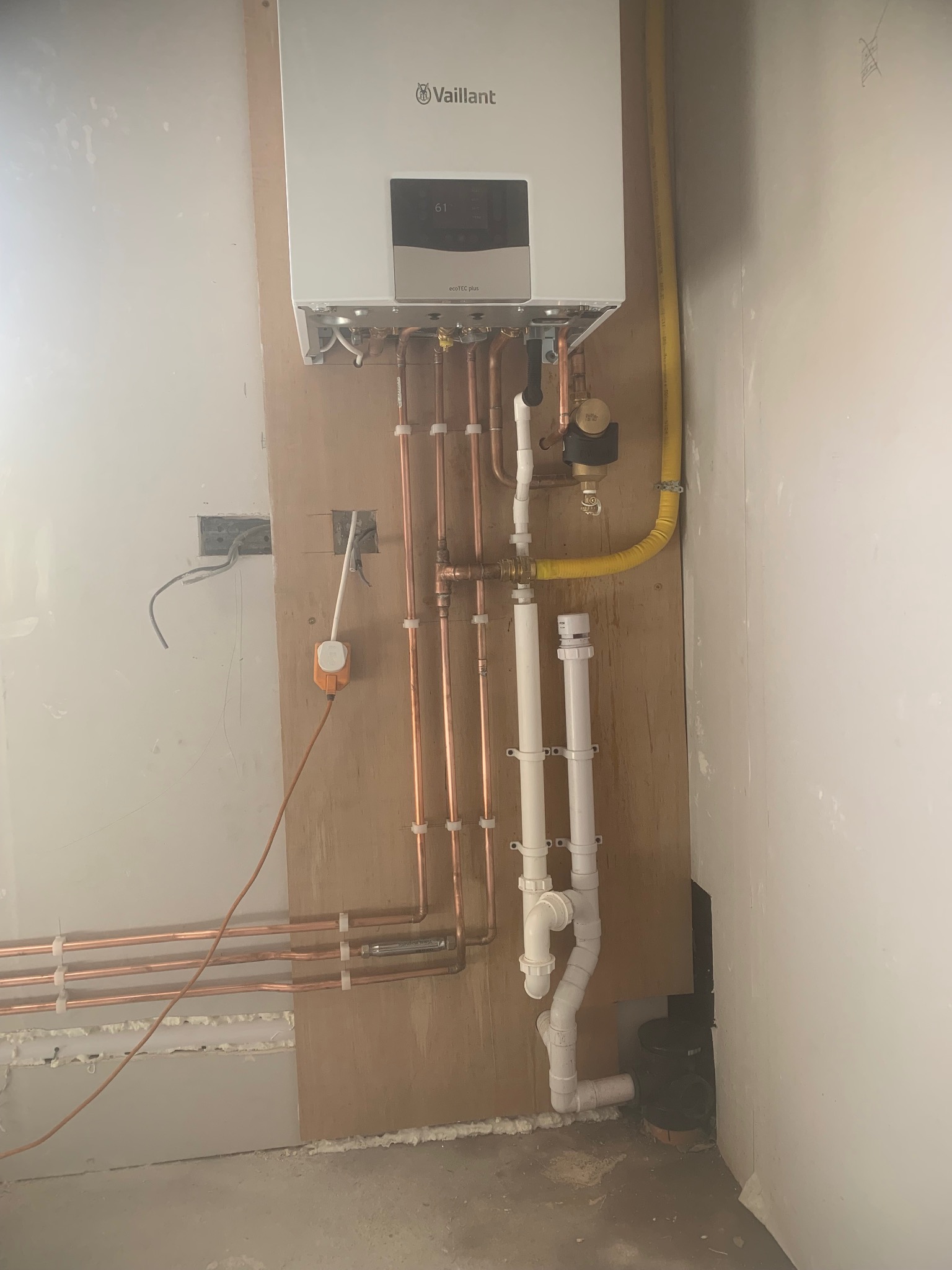 Zamontowany piec gazowy Vaillant z miedzianą instalacją, widoczne rury odpływowe i żółty przewód gazowy. Ściana w trakcie wykończenia, widoczne gniazdka elektryczne.