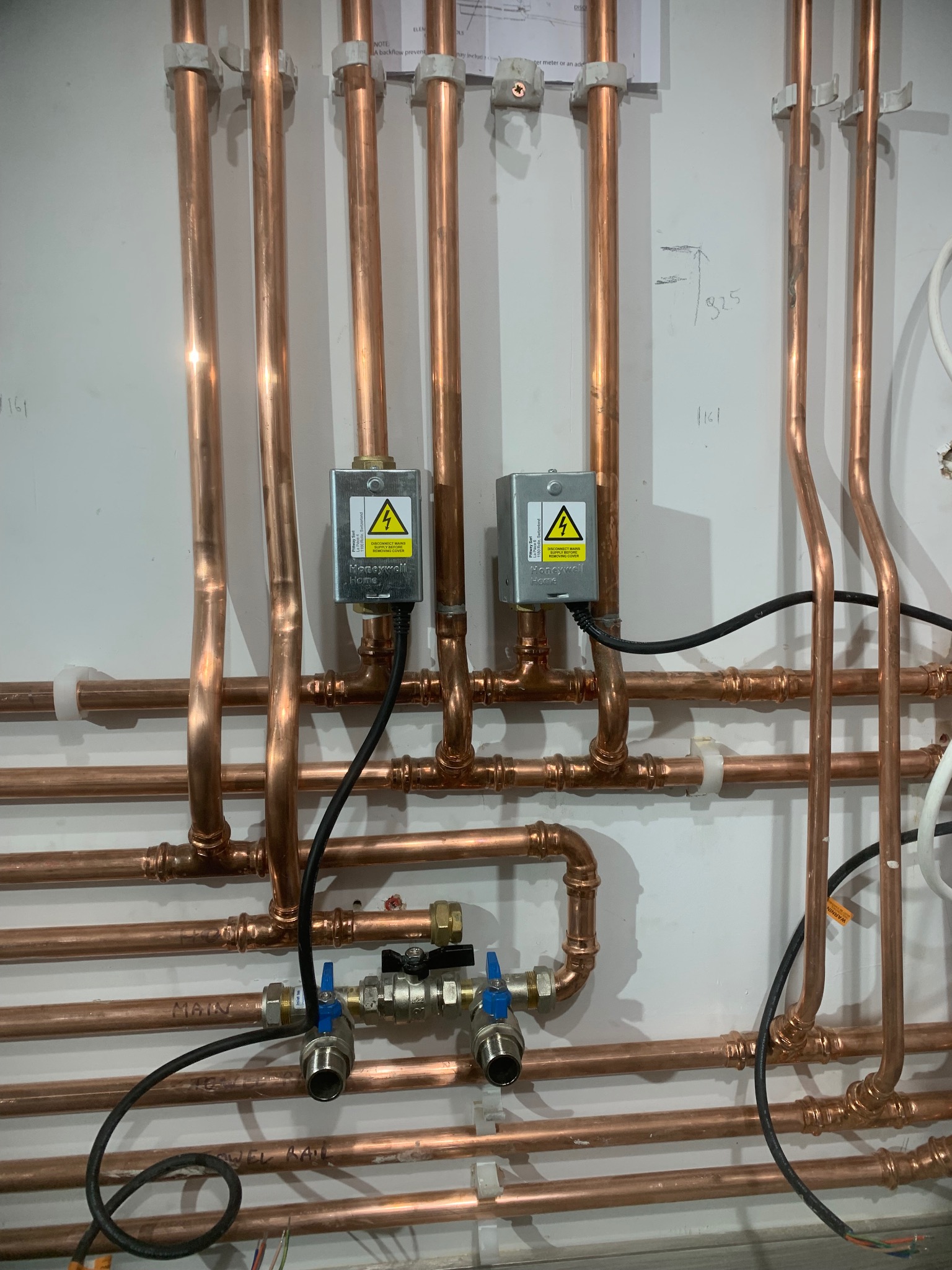 Miedziana instalacja grzewcza z zaworami i elementami elektrycznymi na białej ścianie. Widoczne rury, złączki, przewody i ostrzegawcze etykiety na urządzeniach.