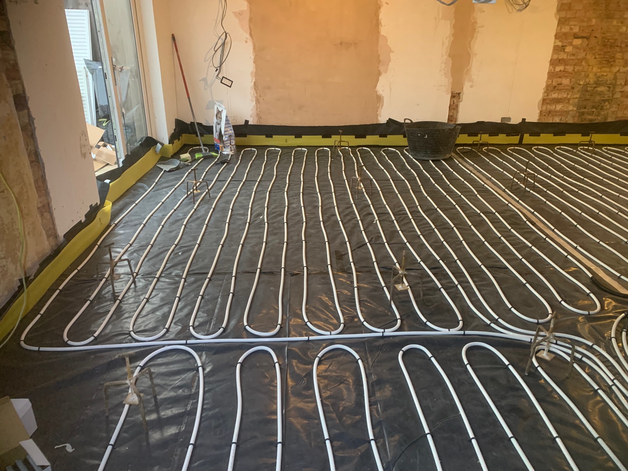 Białe rury ogrzewania podłogowego ułożone na czarnej folii z żółtymi brzegami, widoczne uchwyty mocujące i fragment ściany w trakcie remontu.