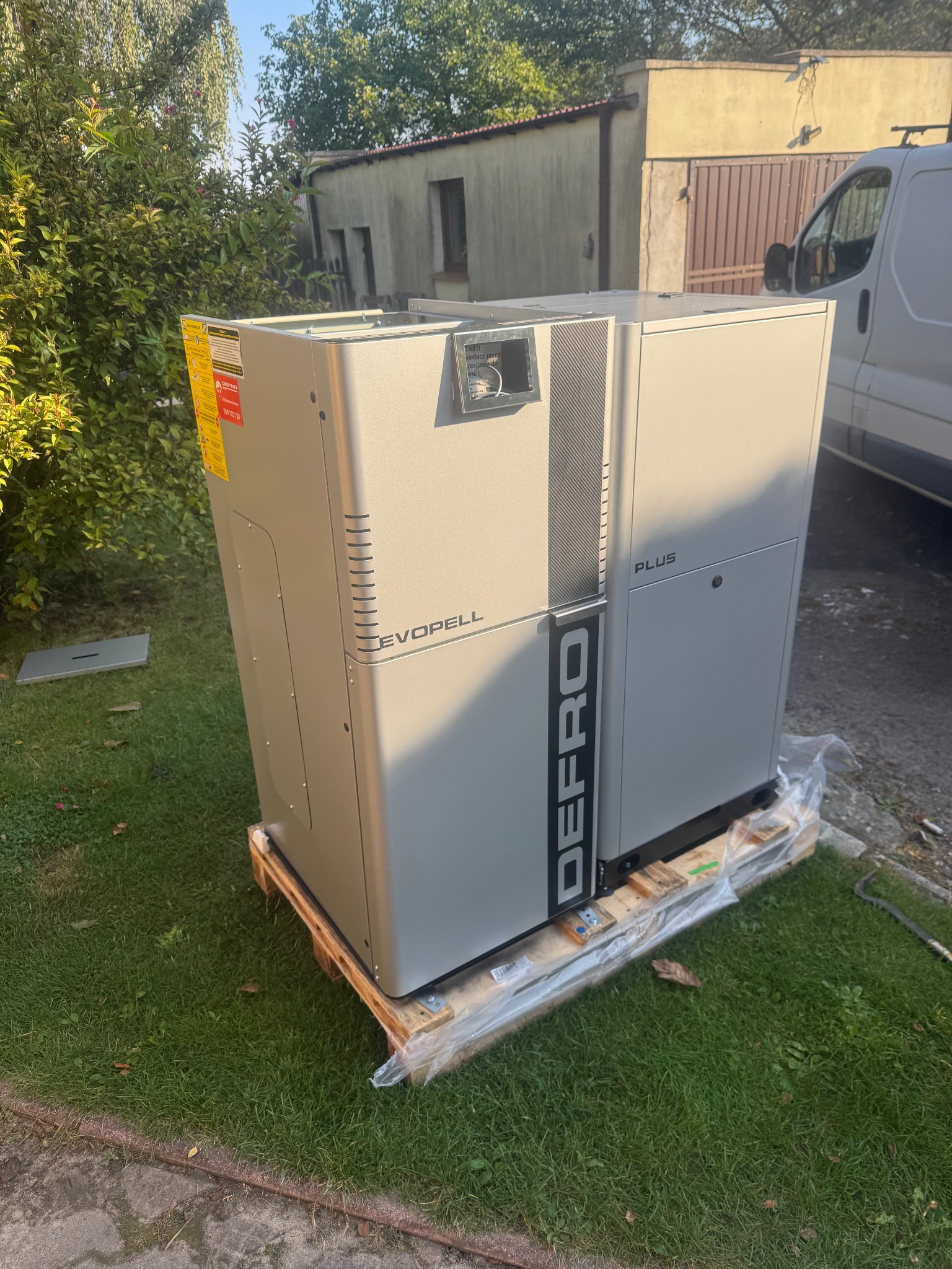 Szara pompa ciepła Defro Evopell Plus na palecie, na trawie, z budynkiem w tle. Widoczny fragment samochodu dostawczego. Słoneczny dzień.