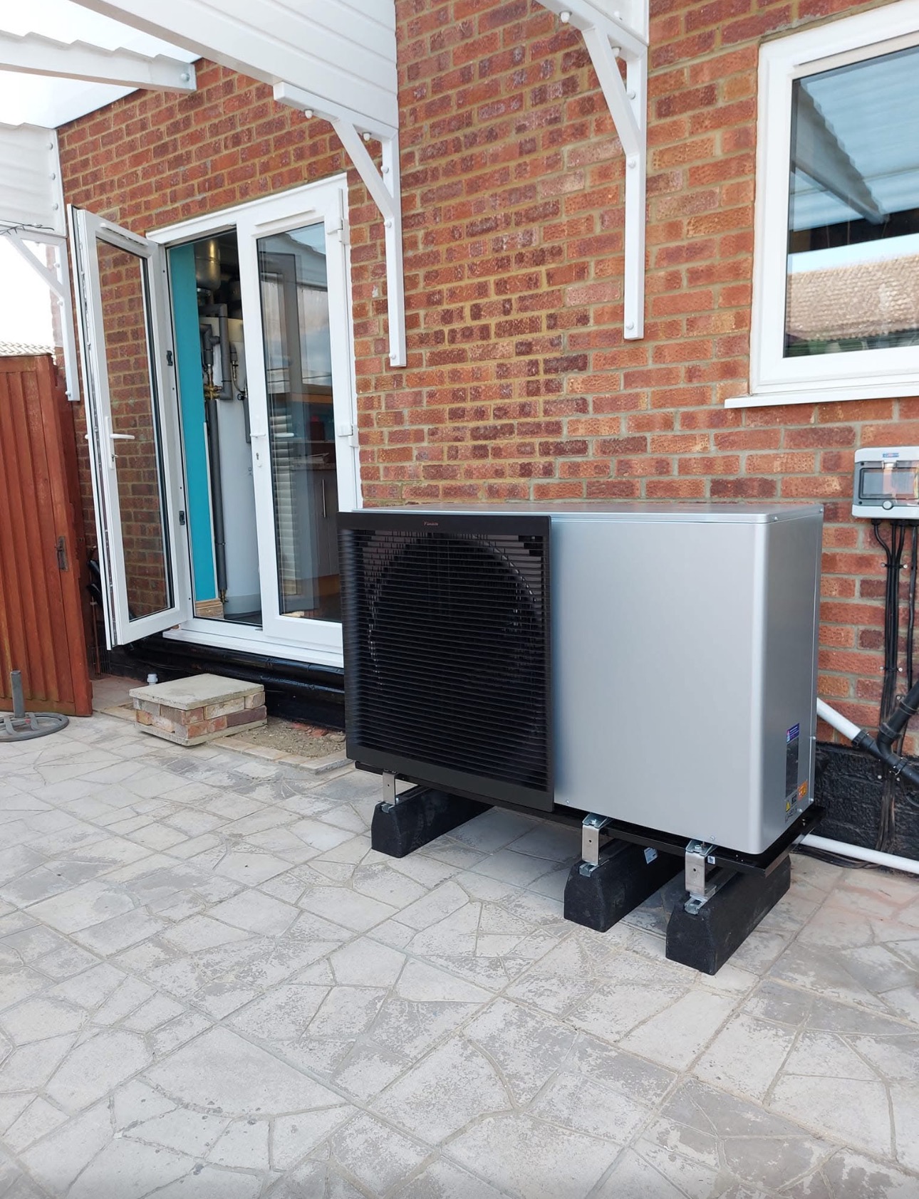 Zewnętrzna jednostka pompy ciepła Daikin Altherma 3 R ECH2O, zamontowana na betonowym patio przy ceglanym budynku z otwartymi drzwiami balkonowymi i oknem.