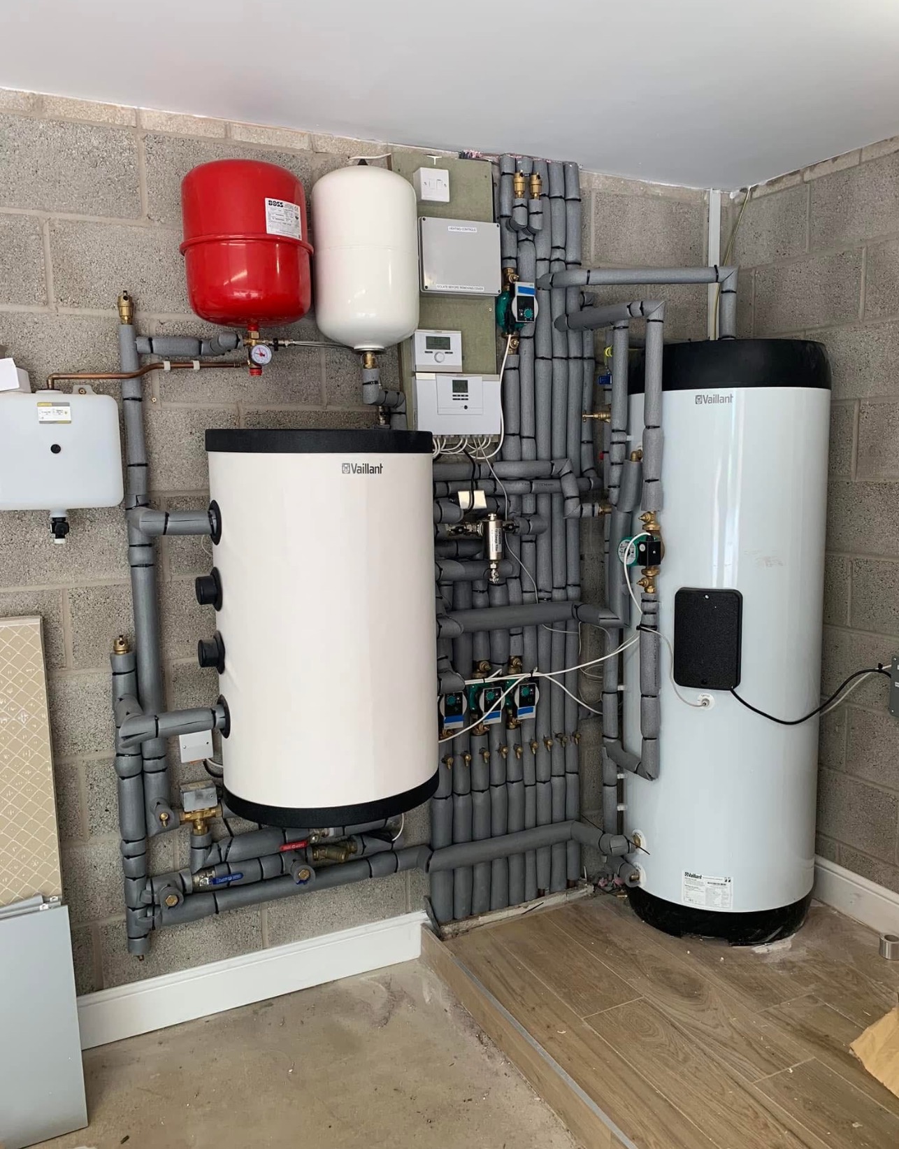 Dwa białe zasobniki Vaillant, czerwony zbiornik wyrównawczy i szare rury izolowane na tle ściany z betonu komórkowego. Instalacja w kotłowni.
