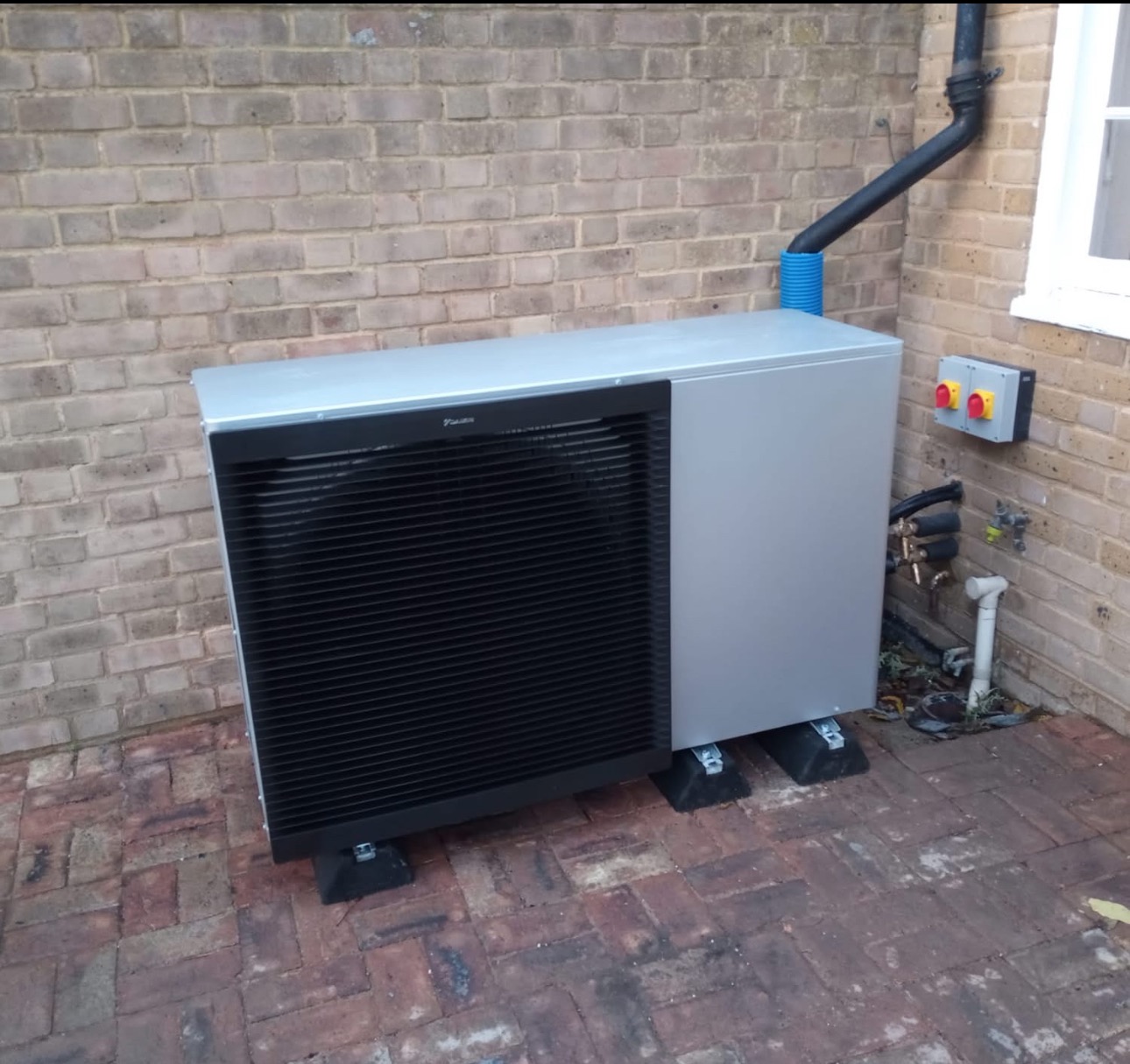 Pompa ciepła Daikin zamontowana na zewnątrz budynku w Lublinie, widoczna na tle ceglanej ściany i kostki brukowej. Instalacja z rurami i skrzynką sterowniczą.