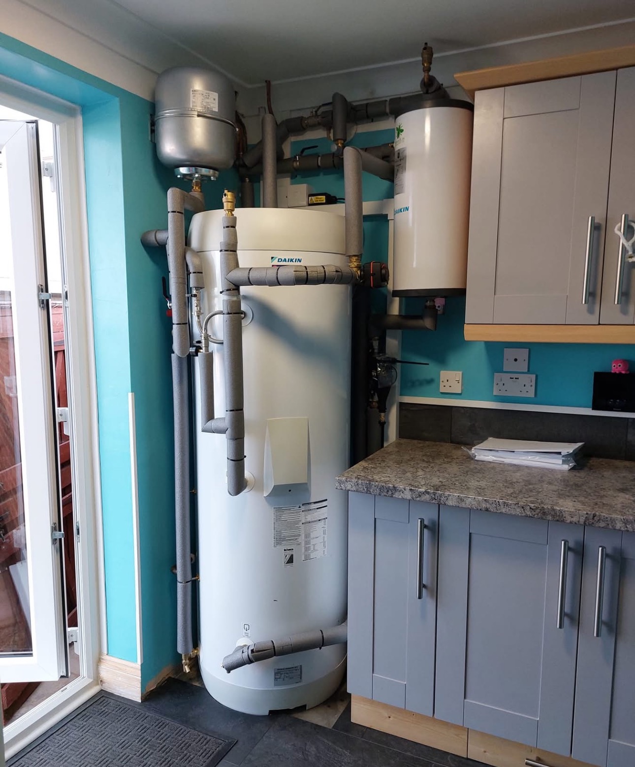 Daikin: Biały zasobnik ciepłej wody użytkowej z szarymi rurami w izolacji, obok szare szafki kuchenne i turkusowa ściana. Widoczny fragment drzwi balkonowych.