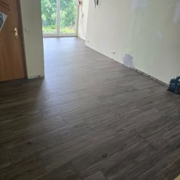 Bartosz Cala - Ułożona, ciemna podłoga z płytek imitujących drewno w jasnym pokoju z oknem balkonowym. Widoczne narzędzia glazurnicze i fragment ściany przygotowanej do malowania.