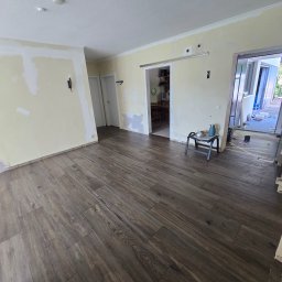 Bartosz Cala - Ułożona podłoga z paneli imitujących drewno w jasnym pokoju z przygotowanymi ścianami do malowania. Widoczne drzwi przesuwne i akcesoria do picia.