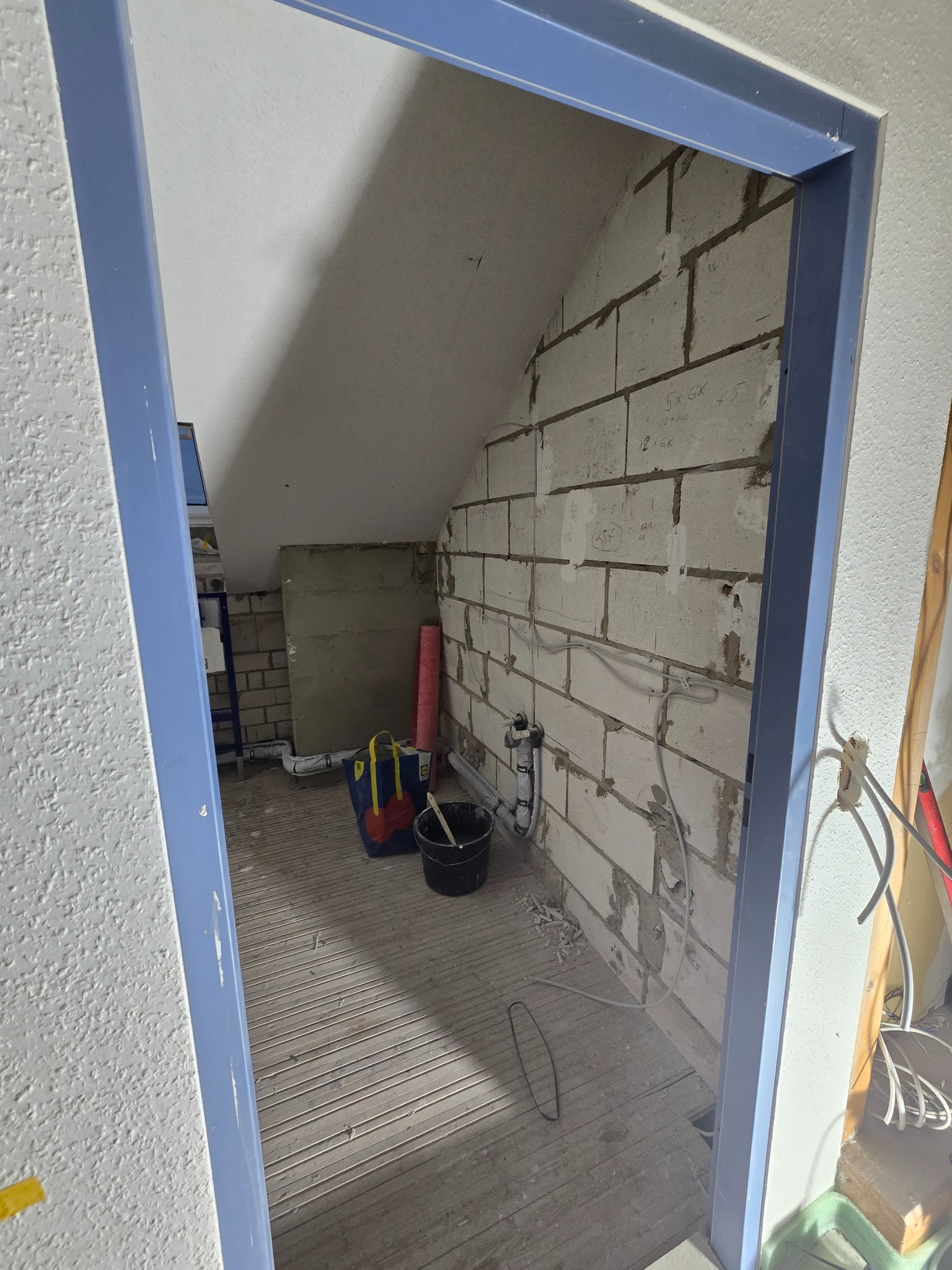 Surowy stan poddasza z widoczną ścianą z bloczków, instalacją i niebieską futryną drzwiową. Przygotowanie do prac wykończeniowych.