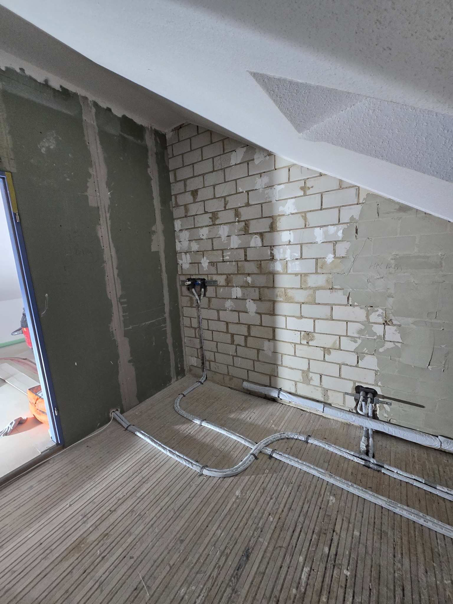 Wnętrze w trakcie prac: ściana z cegły częściowo otynkowana, płyta karton-gips, instalacja podłogowa z rur w izolacji. Widoczne przewody i elementy instalacji.