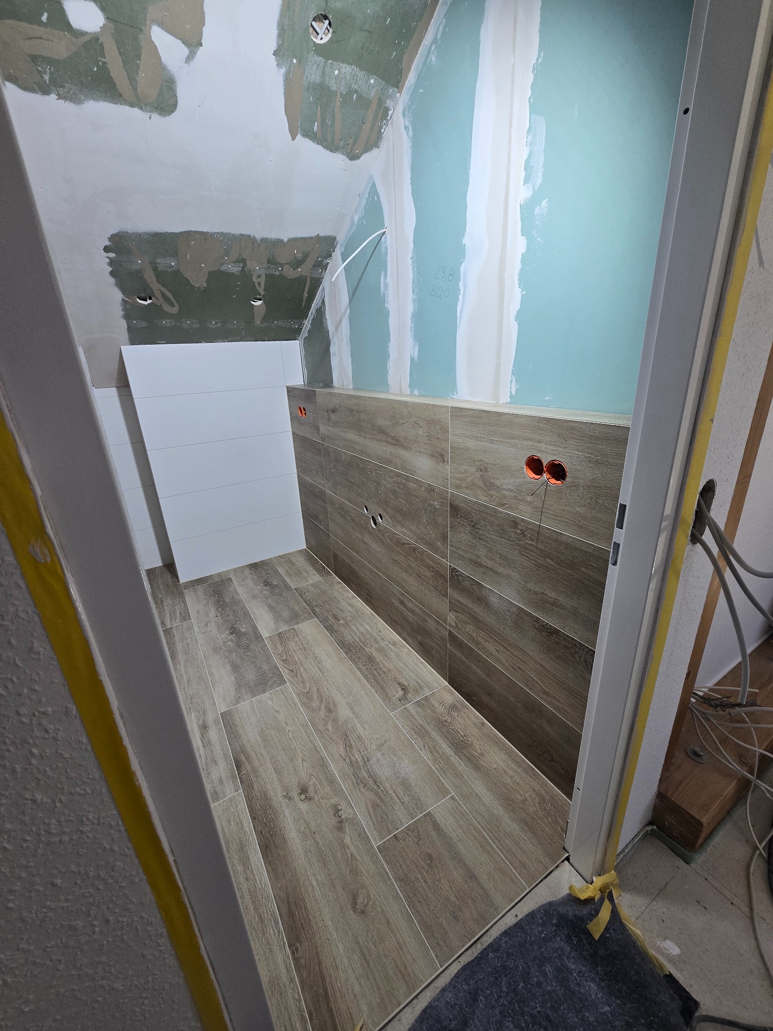 Łazienka w trakcie remontu: ułożone płytki imitujące drewno na podłodze i ścianie, białe płytki na fragmencie ściany, widoczne puszki elektryczne, płyta gipsowa na ścianie.