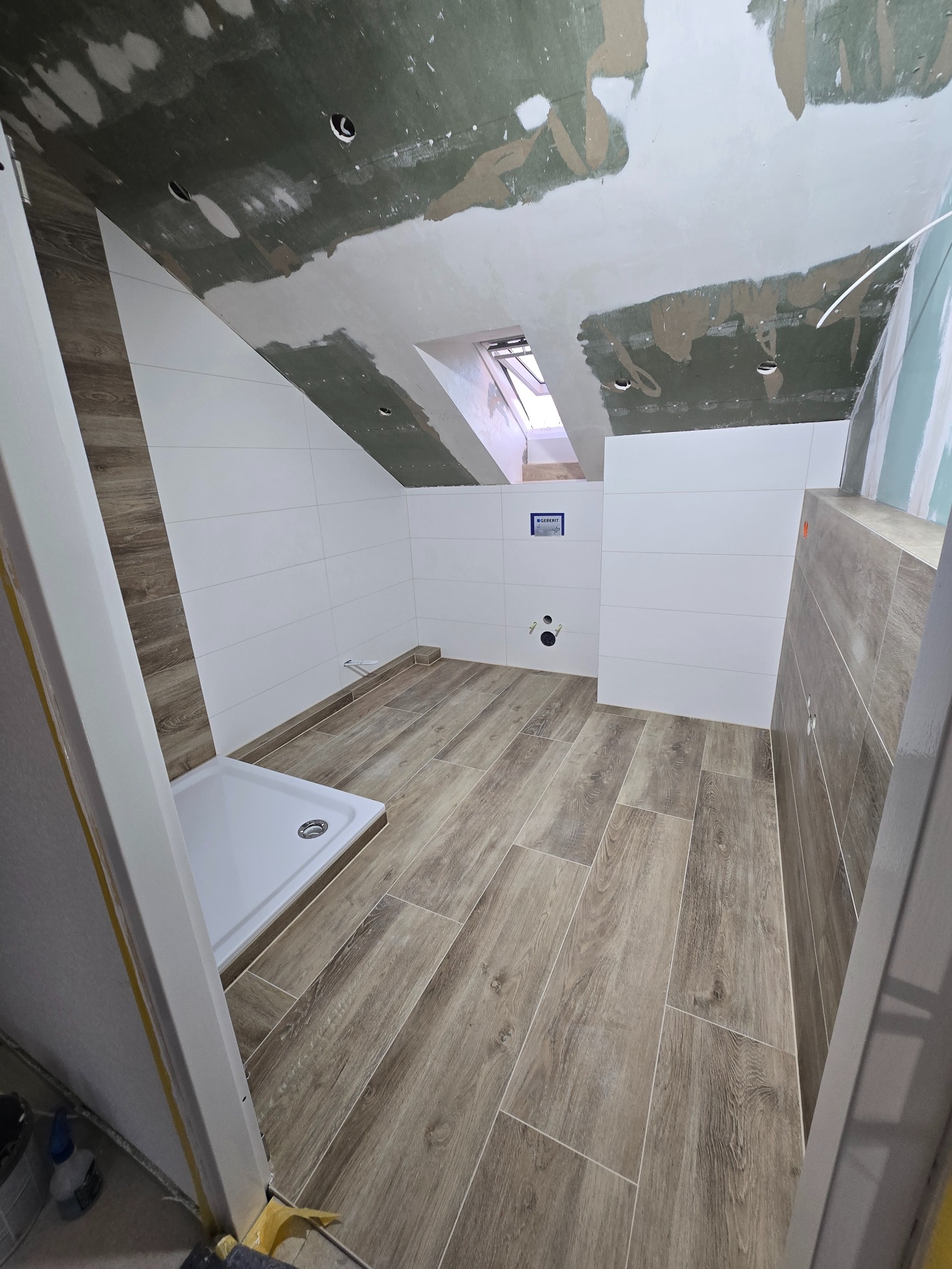 Wykończona łazienka na poddaszu w Mogilnie z drewnopodobną podłogą, białe płytki na ścianach i brodzik. Widoczne okno dachowe i surowy sufit.