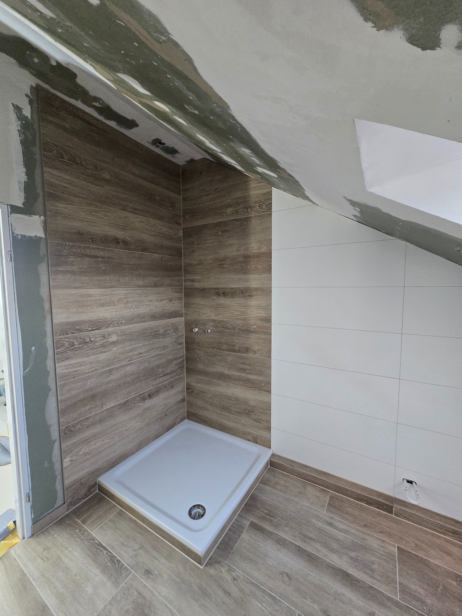 Wykończona kabina prysznicowa z brązowymi płytkami imitującymi drewno i białymi płytkami na ścianie, widoczny brodzik i fragment sufitu w trakcie prac wykończeniowych.