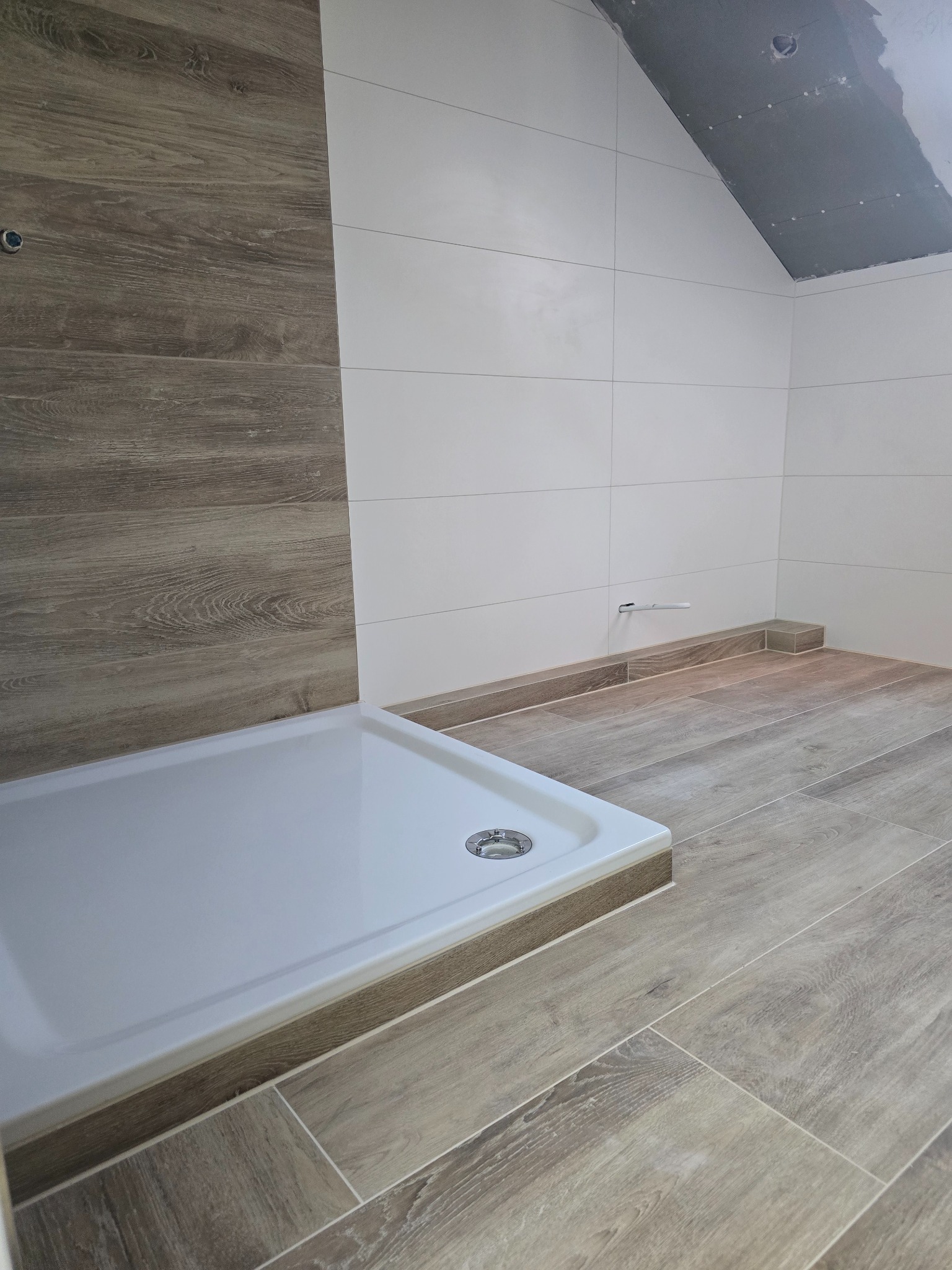 Wykończona łazienka z kabiną prysznicową i płytkami imitującymi drewno na podłodze i ścianie. Białe płytki na ścianach. Poddasze w trakcie wykończenia.