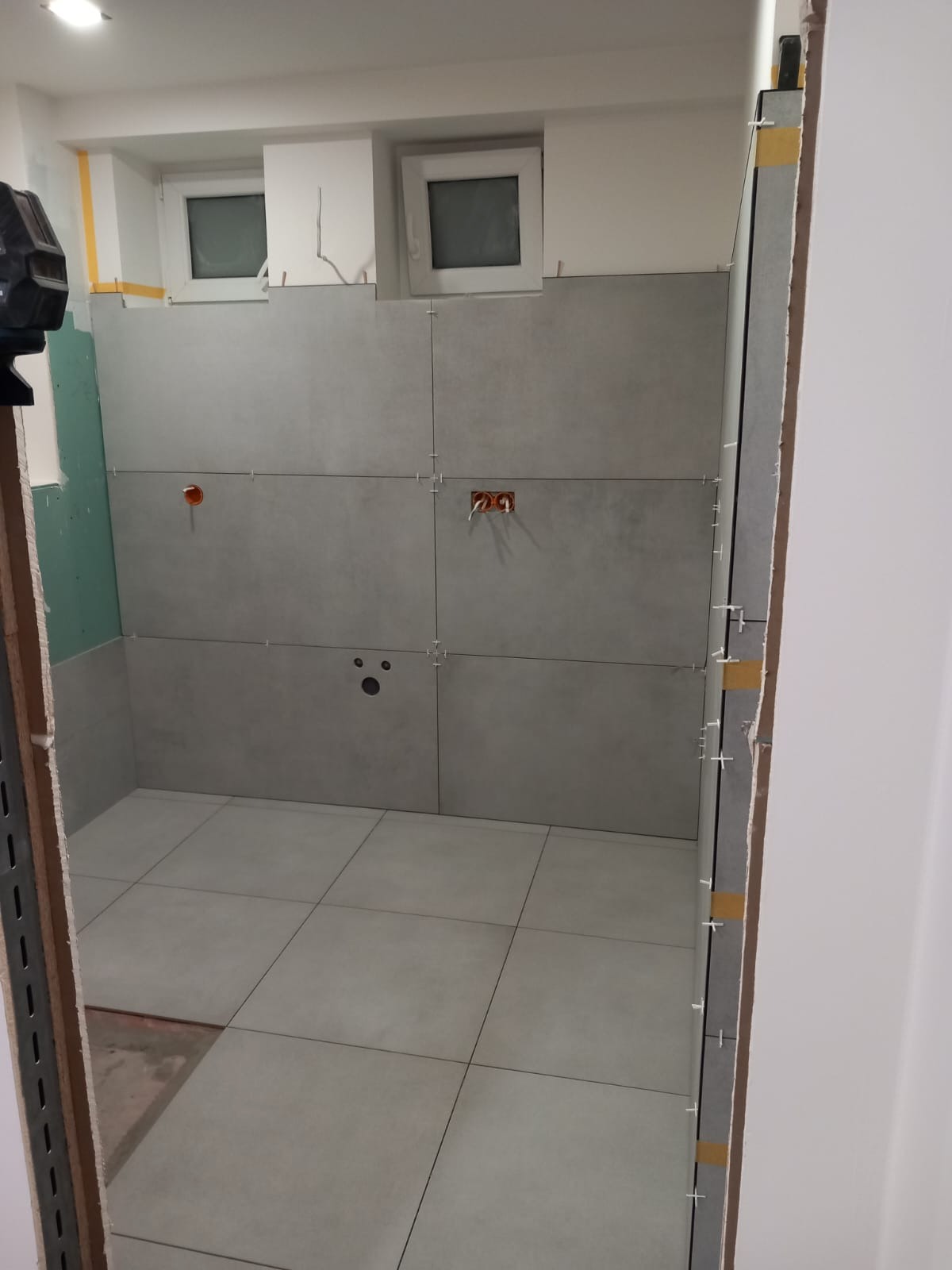 Szare płytki ułożone na ścianach i podłodze w łazience, widoczne krzyżyki dystansowe, przygotowane otwory na instalacje. Ujęcie z perspektywy wejścia.