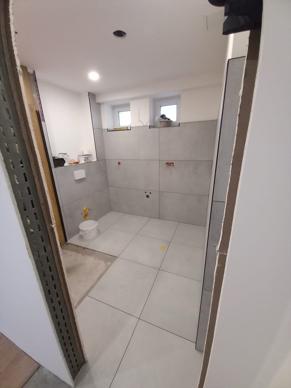 Wnętrze łazienki w trakcie remontu. Widoczne szare płytki na ścianach i podłodze, biała ściana, okno oraz wiadro i narzędzia. Częściowo wykończone pomieszczenie.