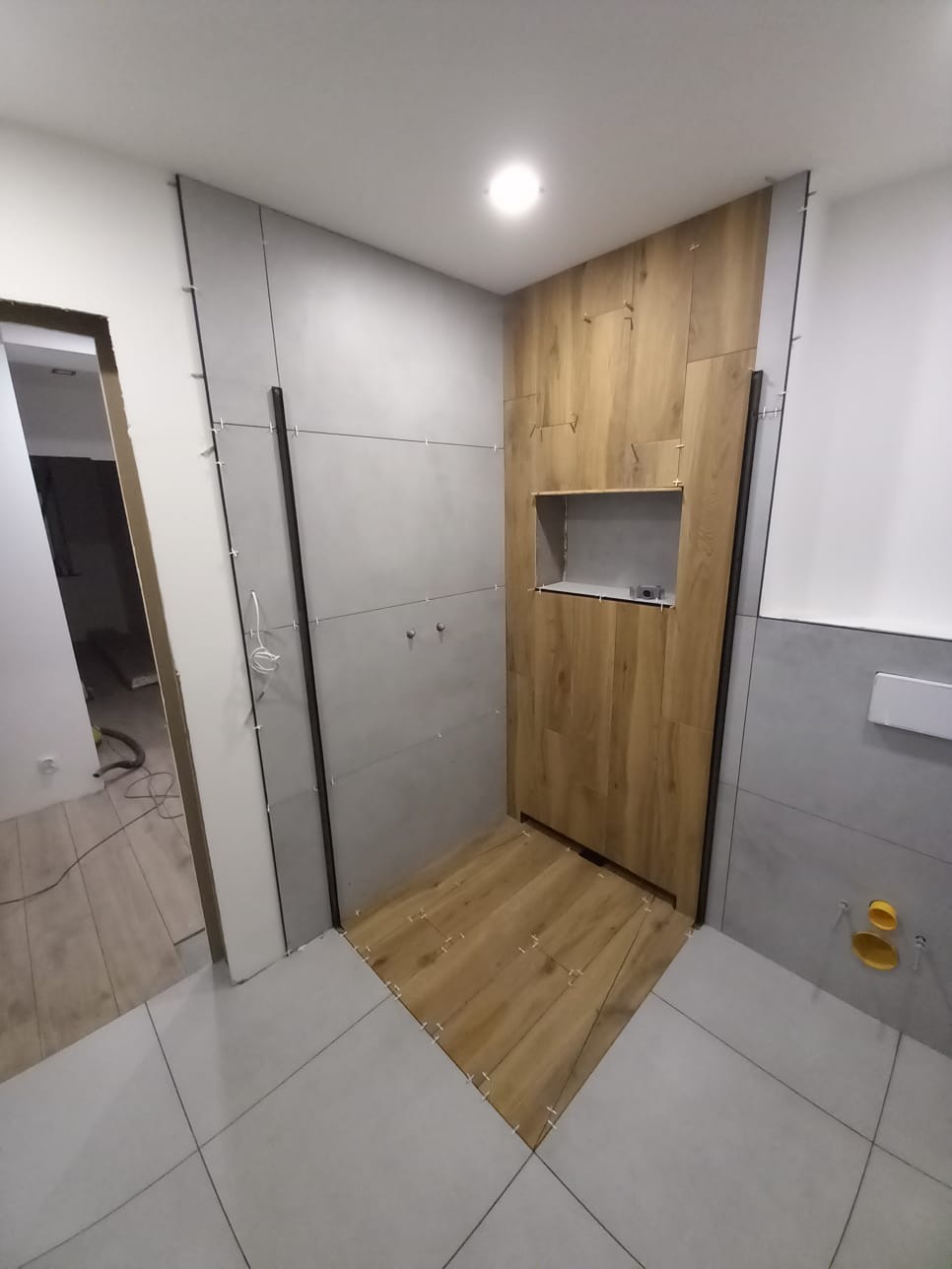 Wnętrze łazienki w trakcie wykańczania: szare płytki ścienne, drewnopodobne płytki podłogowe, widoczne listwy i elementy instalacji. Ukończona wnęka w ścianie.