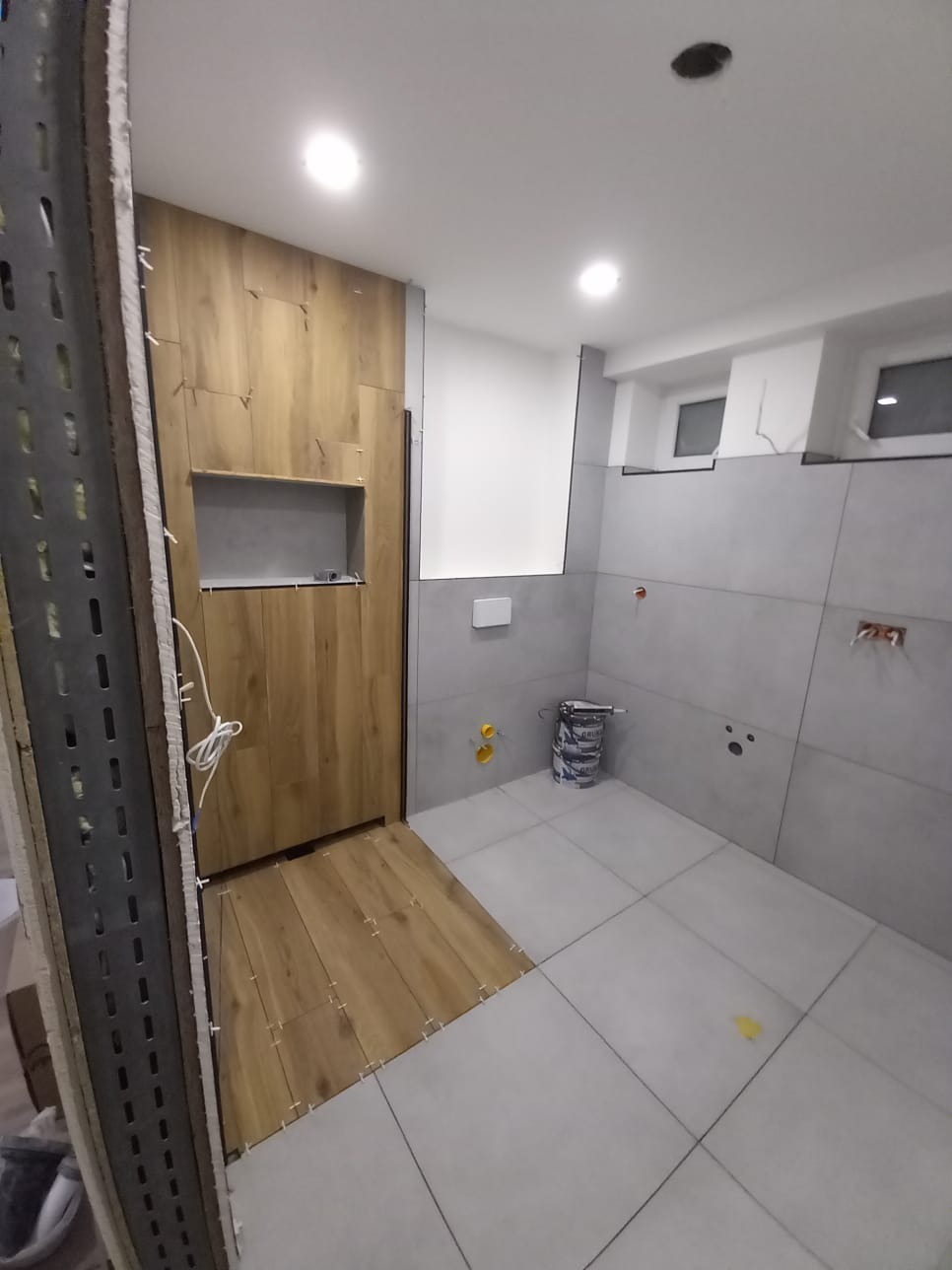 Wnętrze łazienki w trakcie prac: drewnopodobne płytki na ścianie i podłodze, szare płytki na ścianach, widoczne elementy instalacji i wiadro z zaprawą.