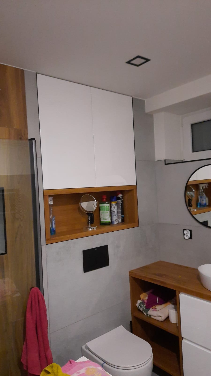 Nowoczesna łazienka z białymi szafkami wiszącymi i drewnianymi elementami. Umywalka z okrągłym lustrem i szare płytki tworzą spójną całość. Widoczna toaleta i akcesoria.