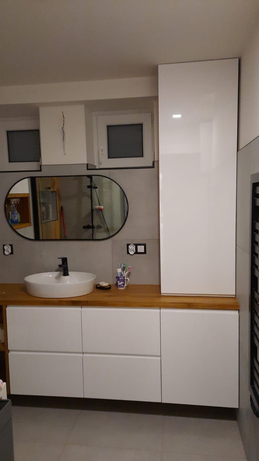 Nowoczesna łazienka z owalnym lustrem, białą umywalką nablatową i drewnianym blatem. Białe szafki pod umywalką i wysoka szafka po prawej stronie. Szare płytki na ścianie.