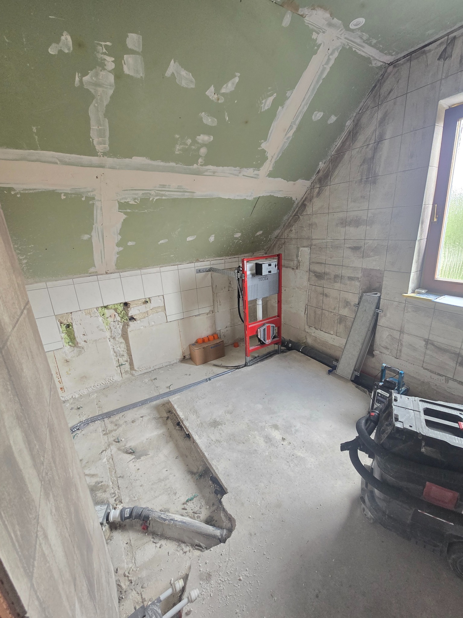 Wnętrze łazienki w trakcie remontu: widoczne instalacje, częściowo położone płytki, przygotowane ściany pod wykończenie, okno z widokiem na zewnątrz.