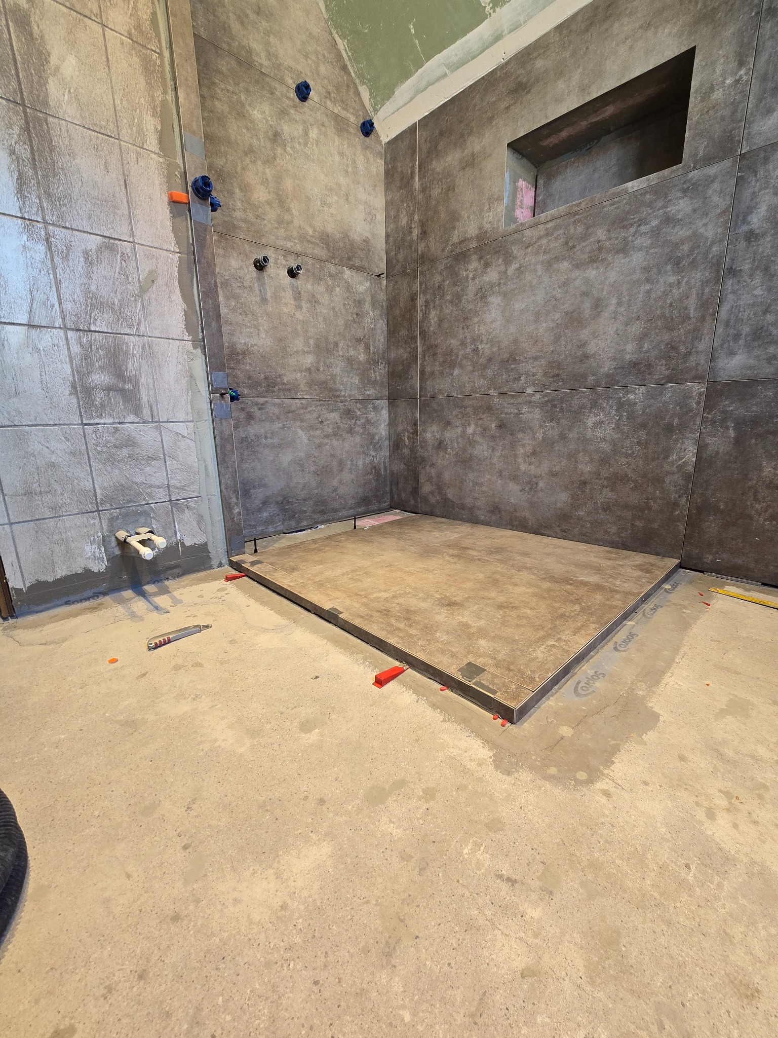 Wnętrze łazienki w trakcie wykańczania. Ułożone płytki podłogowe i ścienne w odcieniach szarości i brązu. Widoczne elementy instalacji i narzędzia glazurnicze.