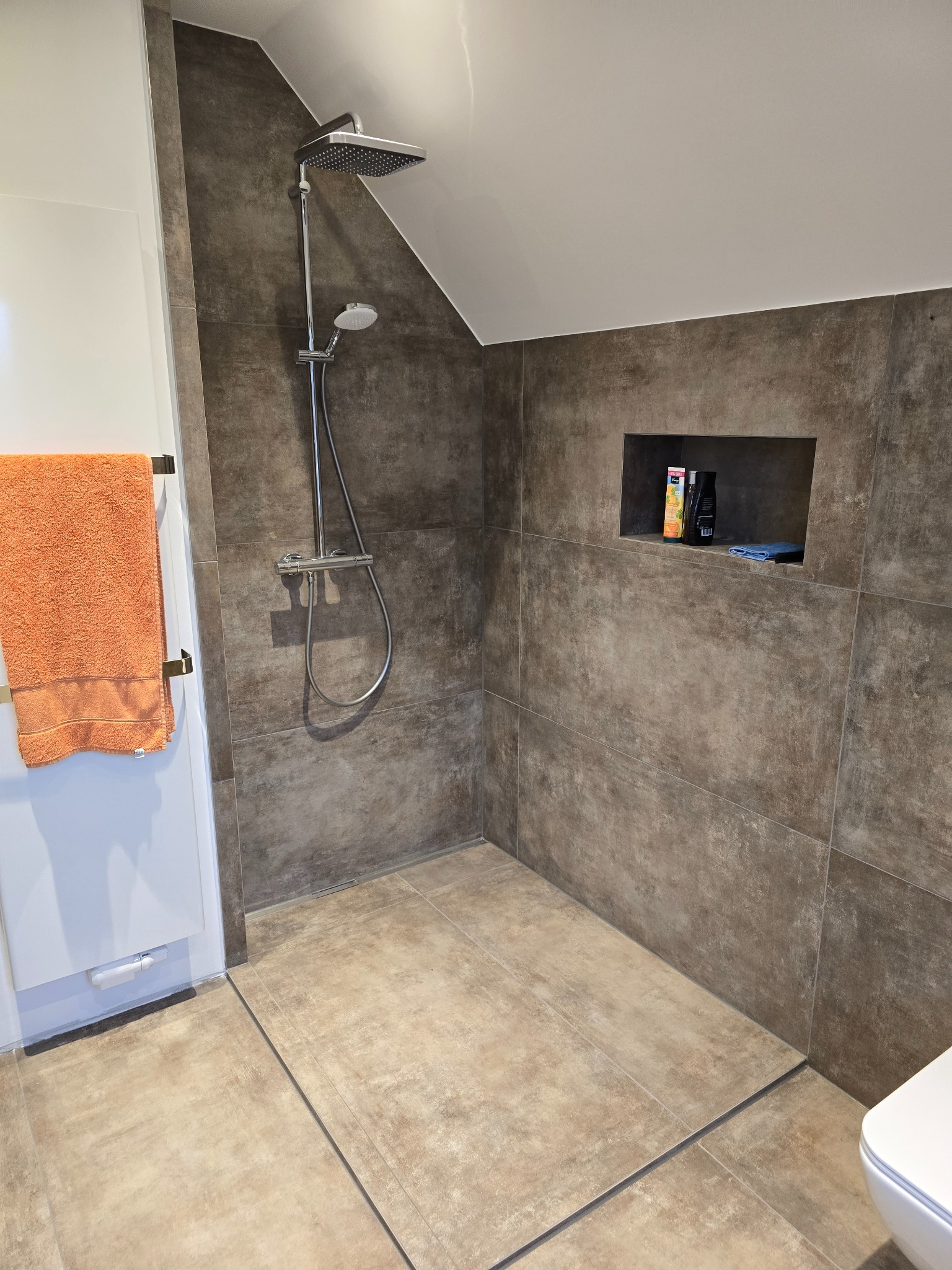 Nowoczesna kabina prysznicowa z brązowymi płytkami, wnęką na kosmetyki i pomarańczowym ręcznikiem. Uwagę zwraca minimalistyczny design i stonowana kolorystyka.