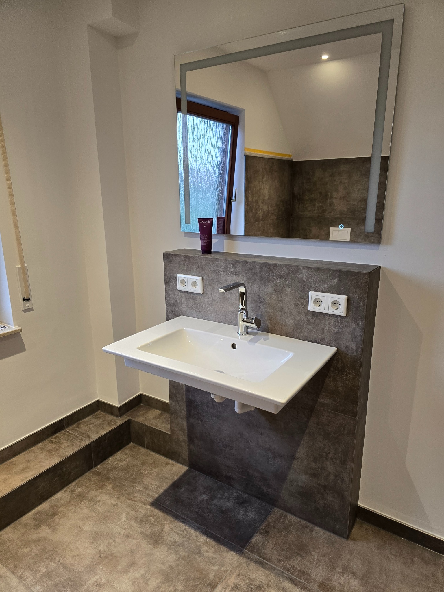 Nowoczesna łazienka z umywalką nablatową na tle grafitowych płytek i podświetlanego lustra. Widoczne gniazdka elektryczne i kosmetyki na blacie.