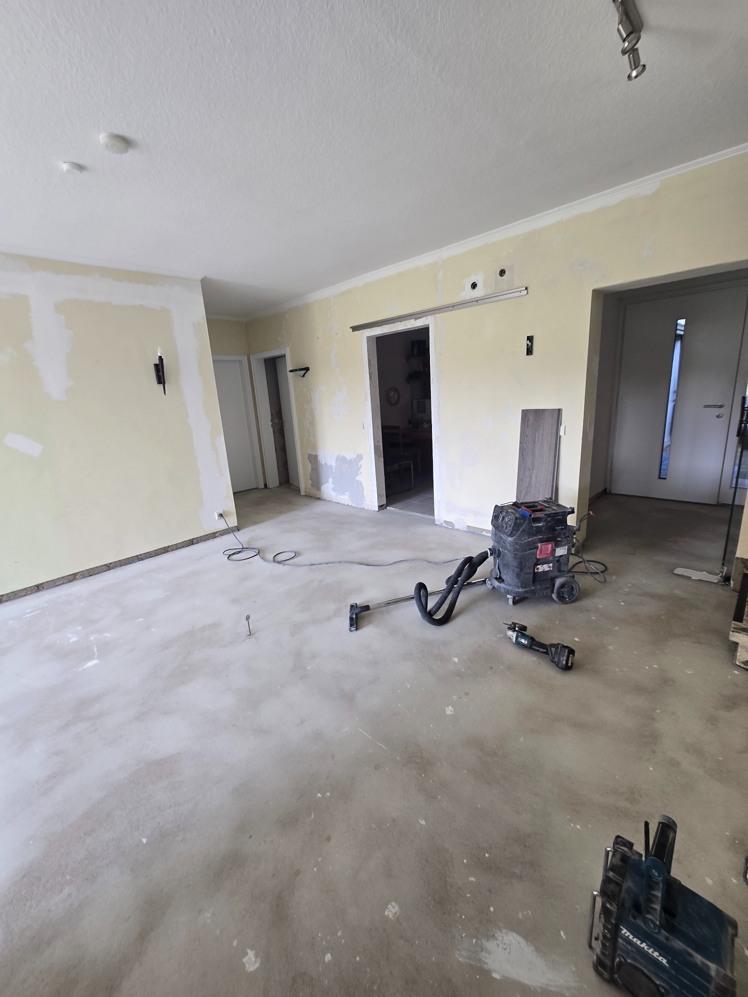 Wnętrze w trakcie remontu: przygotowane ściany do malowania, wylewka na podłodze, widoczny odkurzacz przemysłowy i narzędzia. Etap prac wykończeniowych.