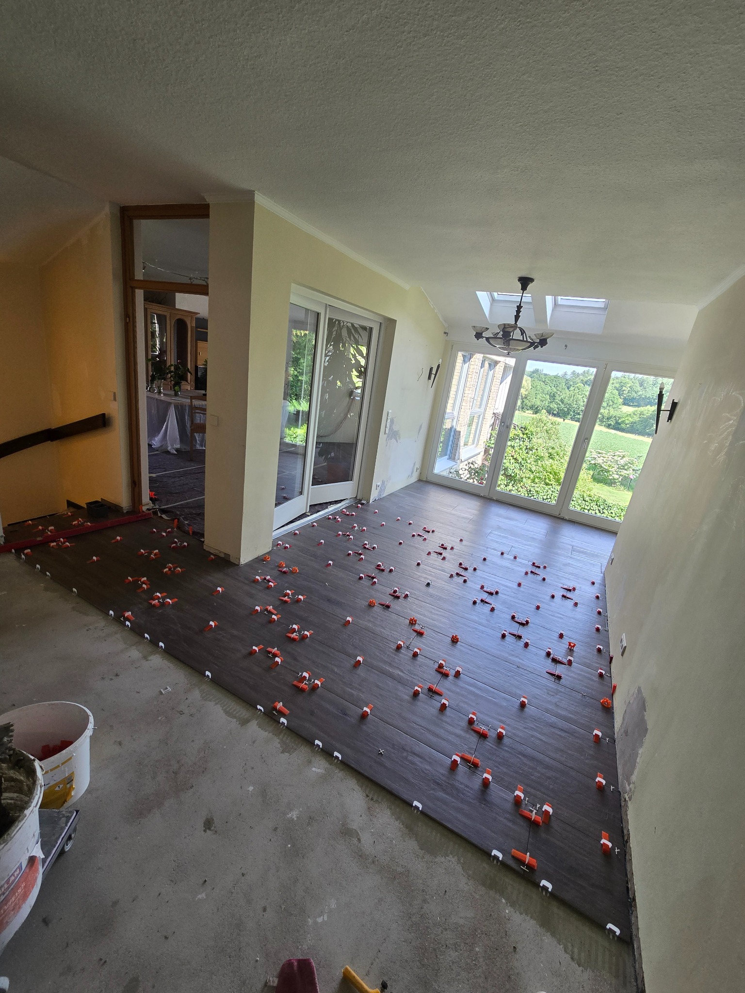 Układanie ciemnych płytek podłogowych z użyciem klipsów poziomujących w przestronnym wnętrzu z dużymi oknami i widokiem na zieleń. Częściowo wykończone podłoże.