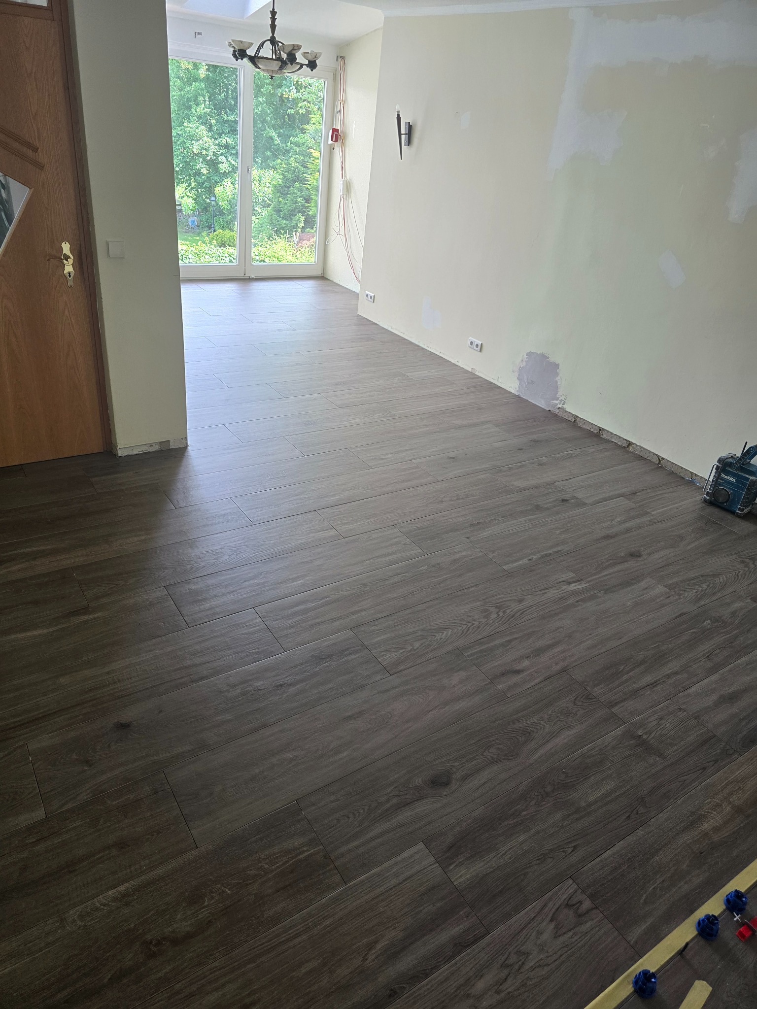 Ułożona, ciemna podłoga z płytek imitujących drewno w jasnym pokoju z oknem balkonowym. Widoczne narzędzia glazurnicze i fragment ściany przygotowanej do malowania.