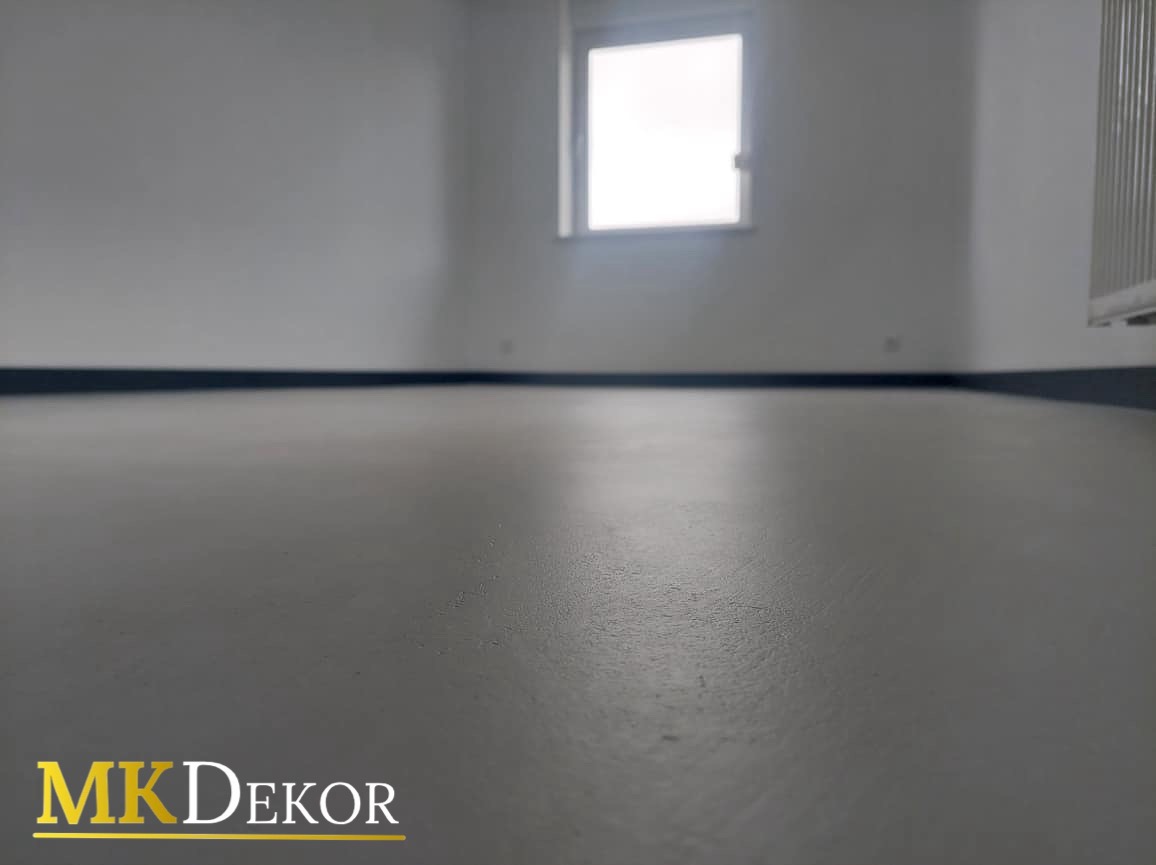 Szara, gładka posadzka w pustym pomieszczeniu z oknem i grzejnikiem. Widoczny fragment listwy przypodłogowej. Logo MK Dekor w lewym dolnym rogu.