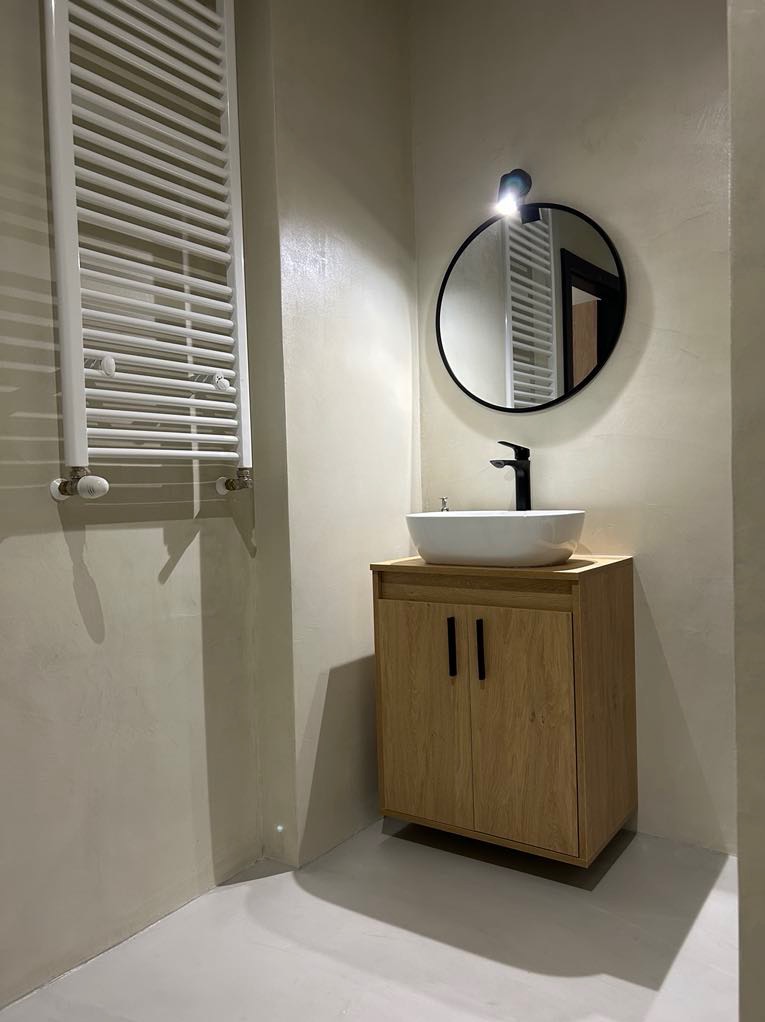 Wykończona łazienka z umywalką nablatową, szafką z jasnego drewna i grzejnikiem drabinkowym. Ściany pokryte dekoracyjnym tynkiem w odcieniach beżu i szarości.