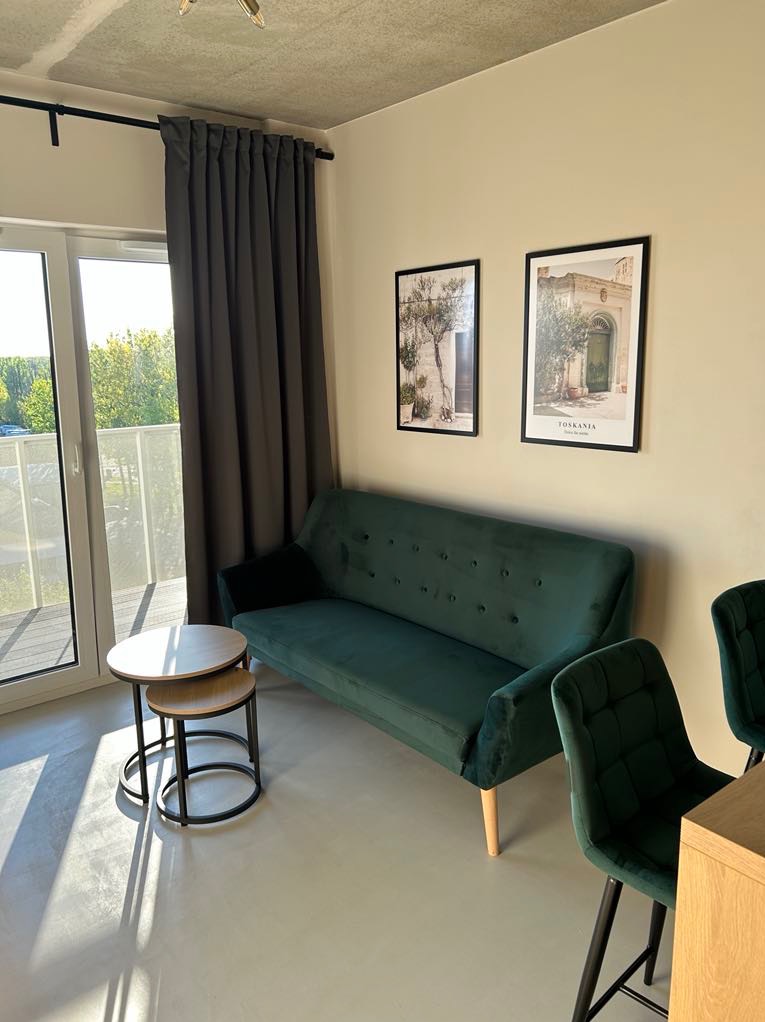 Nowoczesny salon z zieloną sofą i krzesłami, jasną posadzką, plakatami na ścianie i balkonem z widokiem na zieleń. Minimalistyczny design z akcentami kolorystycznymi.