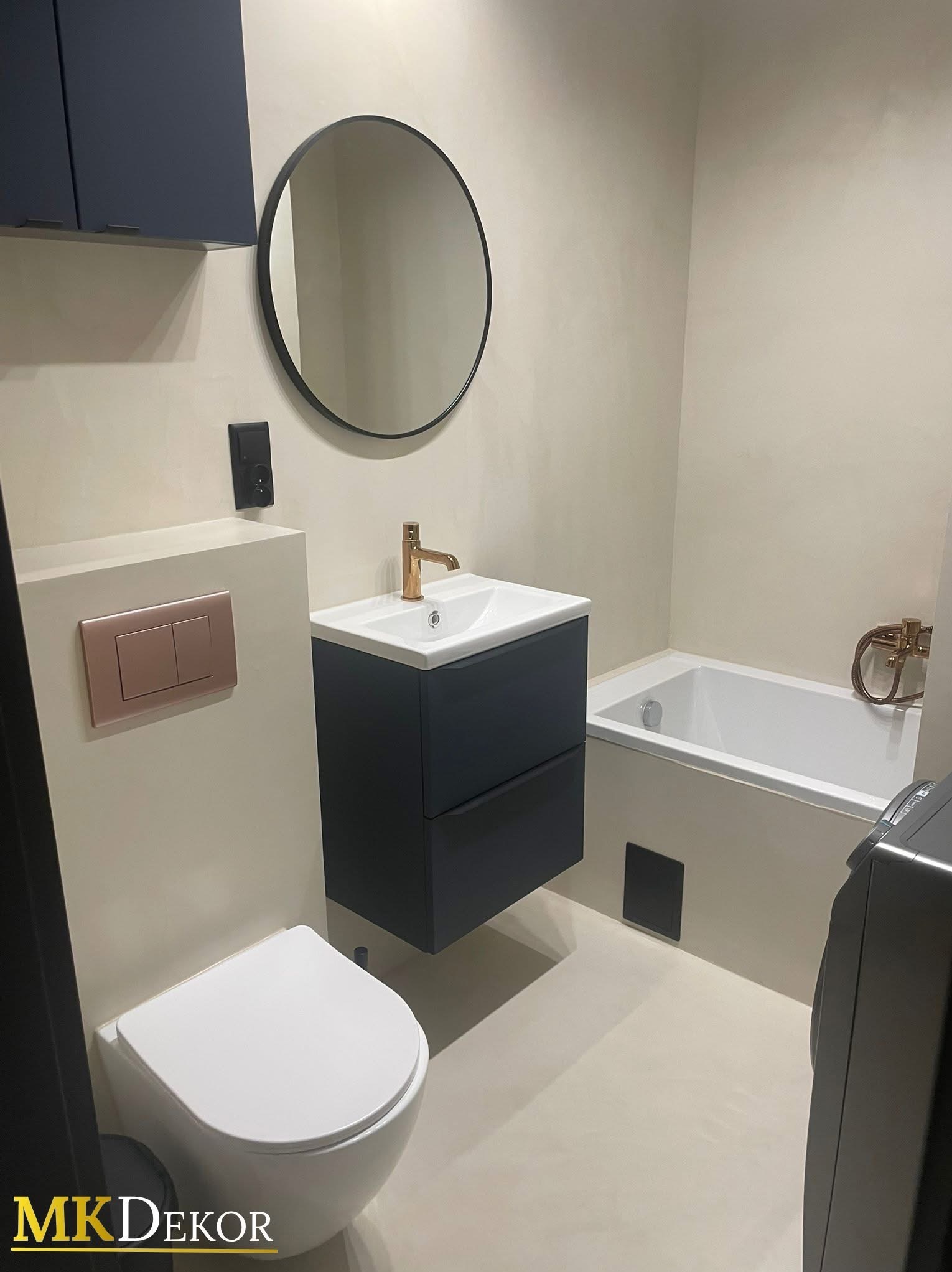 Wykończona łazienka w beżowych odcieniach z umywalką na szafce w kolorze granatowym, okrągłym lustrem i złotą armaturą. Ściany wykończone mikrocementem.