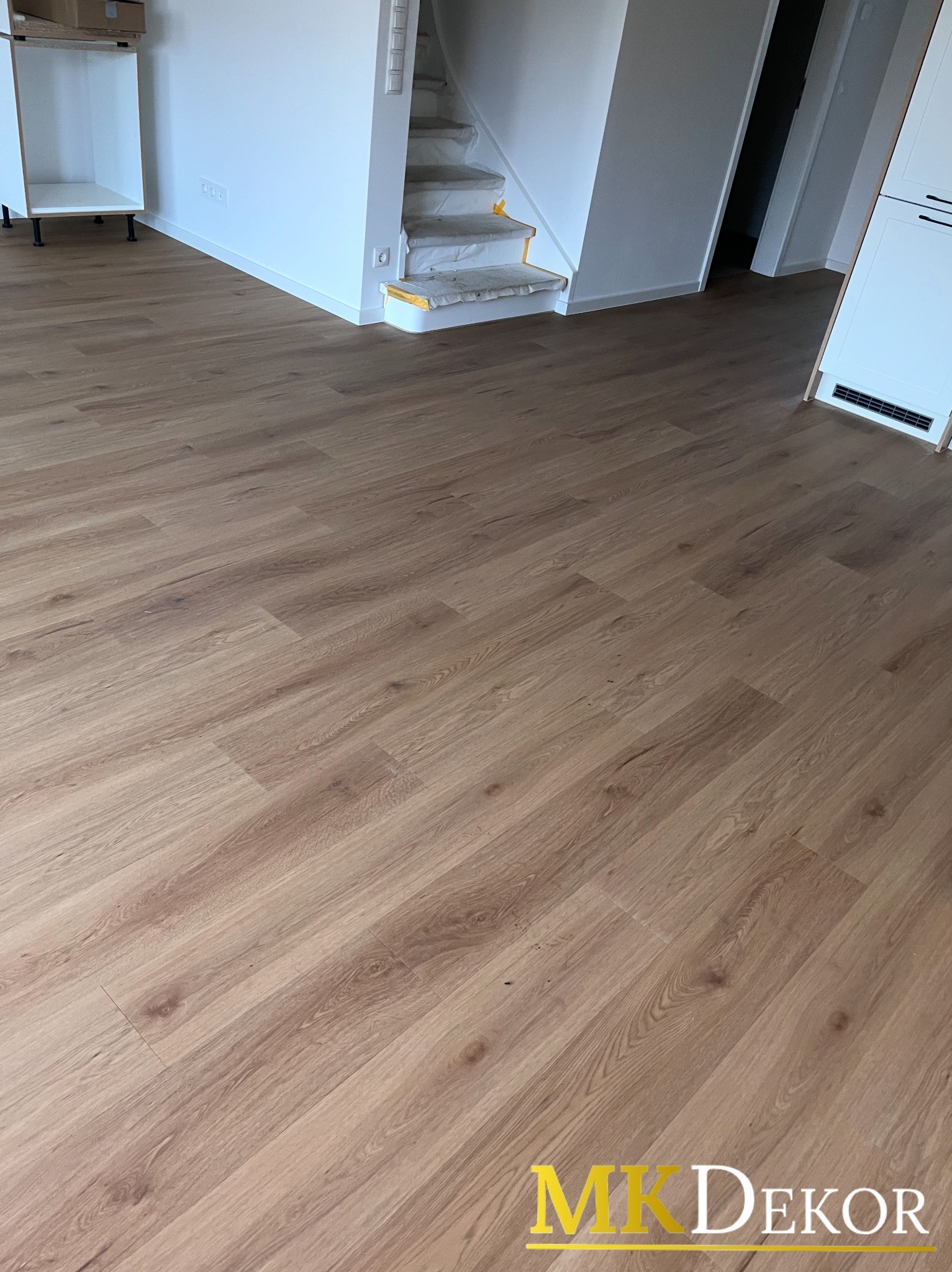 Jasne panele podłogowe ułożone w jodełkę, widoczne fragmenty schodów zabezpieczonych folią i mebli w surowym stanie. Nowoczesne wnętrze w trakcie wykończenia.