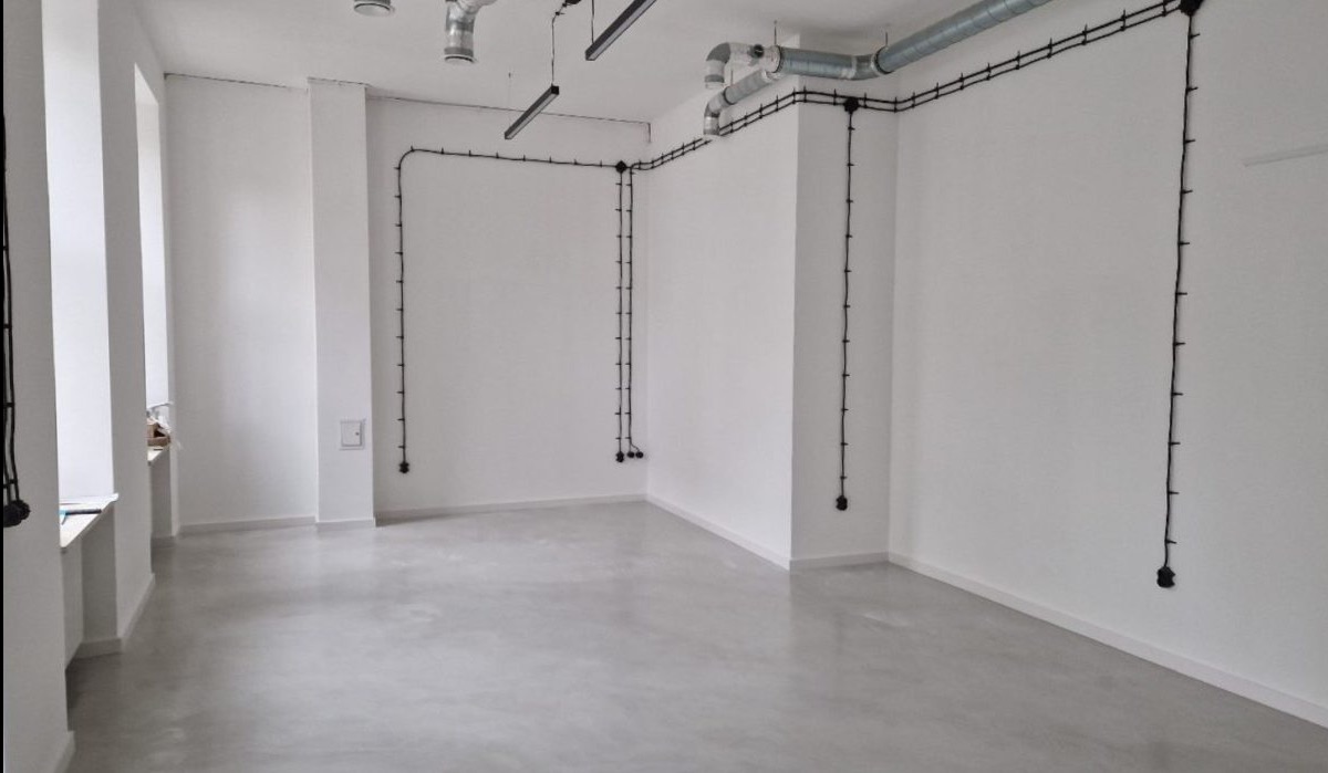 Jasnoszara, gładka posadzka w minimalistycznym wnętrzu z białymi ścianami i widoczną instalacją elektryczną. Surowy, nowoczesny design.