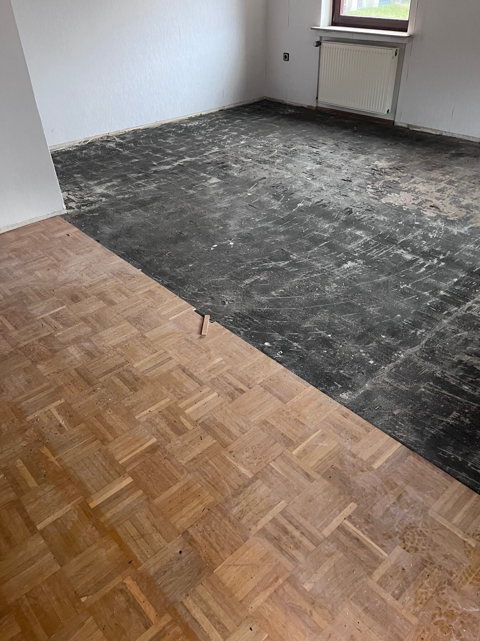 Fragment pokoju z częściowo odnowioną podłogą parkietową w jodełkę. Widoczny kontrast między starą, zszarzałym parkietem a odnowioną, jasną częścią.