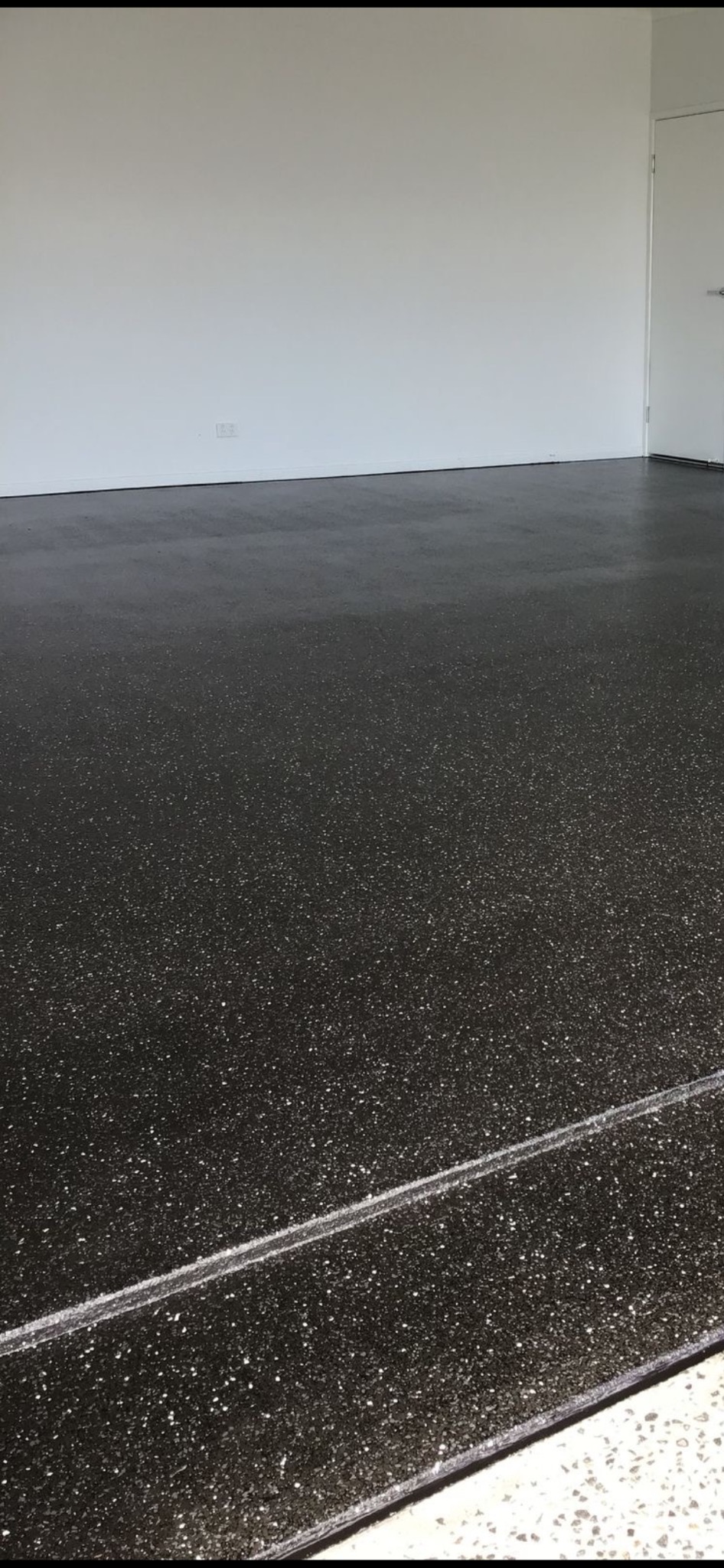 Błyszcząca, ciemna posadzka z drobnymi, jasnymi elementami, świeżo wylana w pustym pomieszczeniu z białą ścianą i drzwiami. Widoczny detal wykończenia przy ścianie.