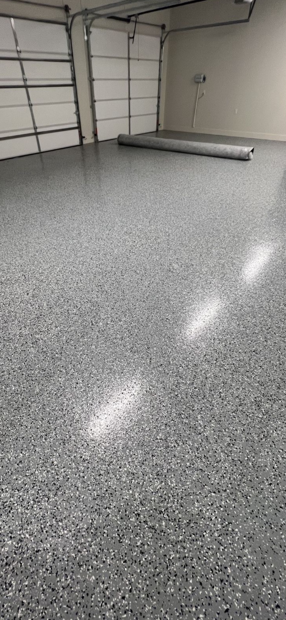 Szara posadzka żywiczna z czarnymi i białymi drobinkami w garażu, z widocznymi drzwiami garażowymi i zwiniętym dywanem w tle. Błyszcząca powierzchnia.