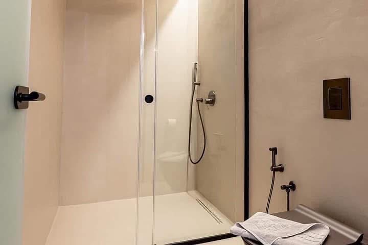 Nowoczesna kabina prysznicowa z mikrocementu w beżowej tonacji, czarne detale armatury, minimalistyczny design, widok na bidet i ręcznik.