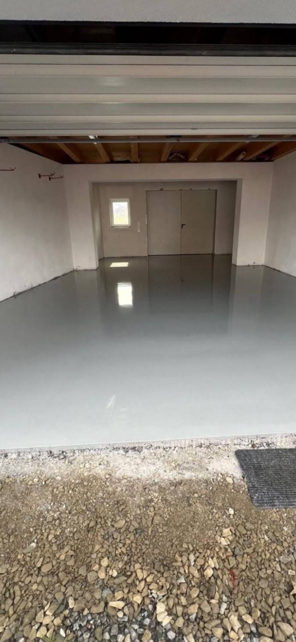Gładka, szara posadzka żywiczna w garażu z widocznym oknem i drzwiami. Podłoże przed wjazdem wyłożone kamieniami. Widoczne elementy konstrukcji dachu.