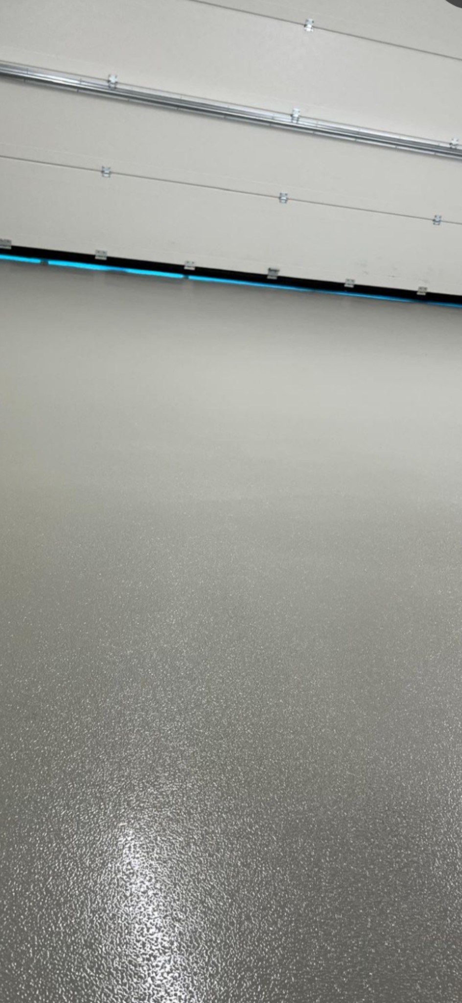 Gładka, szara posadzka z połyskiem, widoczna w dolnej części kadru. W górnej części biała brama garażowa z niebieskim oświetleniem LED. Minimalistyczne ujęcie.