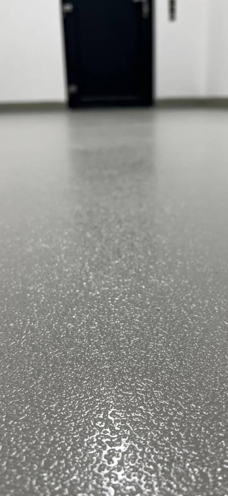 Błyszcząca, szara posadzka z efektem delikatnej struktury, odbijająca światło. W tle widoczny fragment ściany i czarne drzwi.