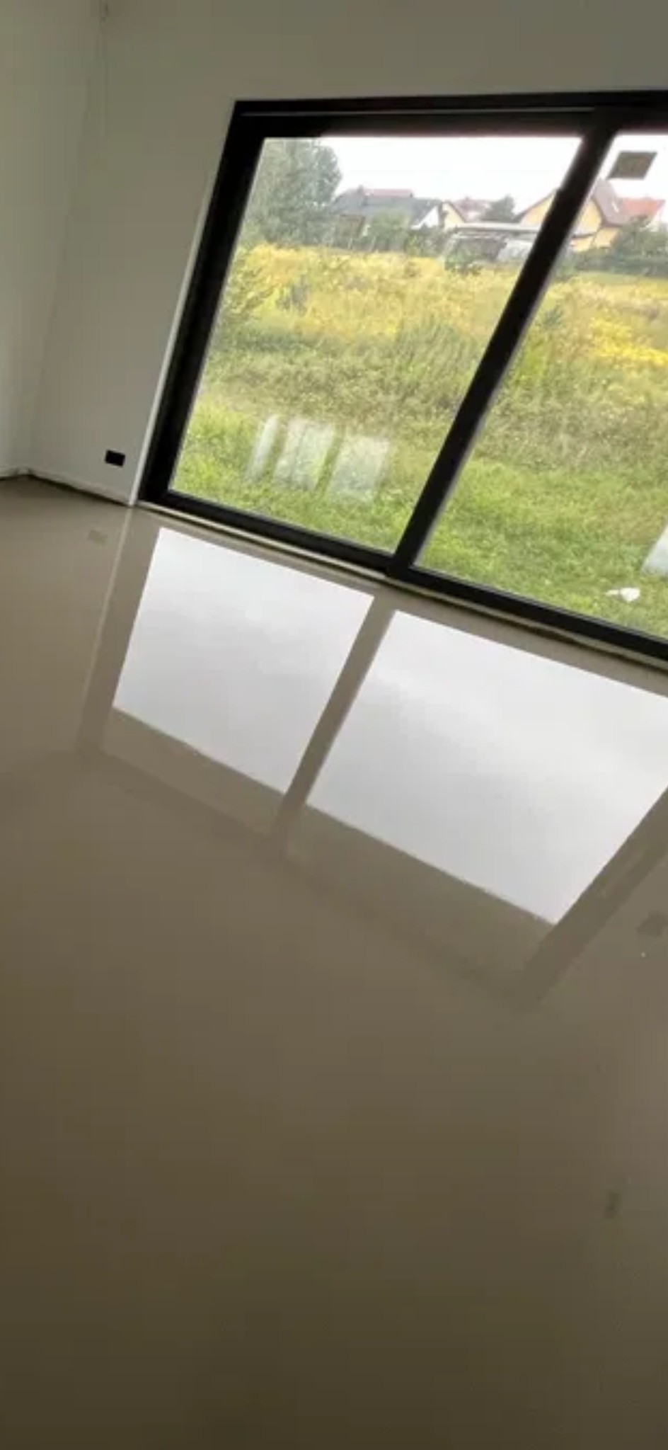 Beżowa, błyszcząca posadzka w nowym wnętrzu z dużym oknem. Widok na zewnątrz odbija się w gładkiej powierzchni podłogi. Minimalistyczny design.