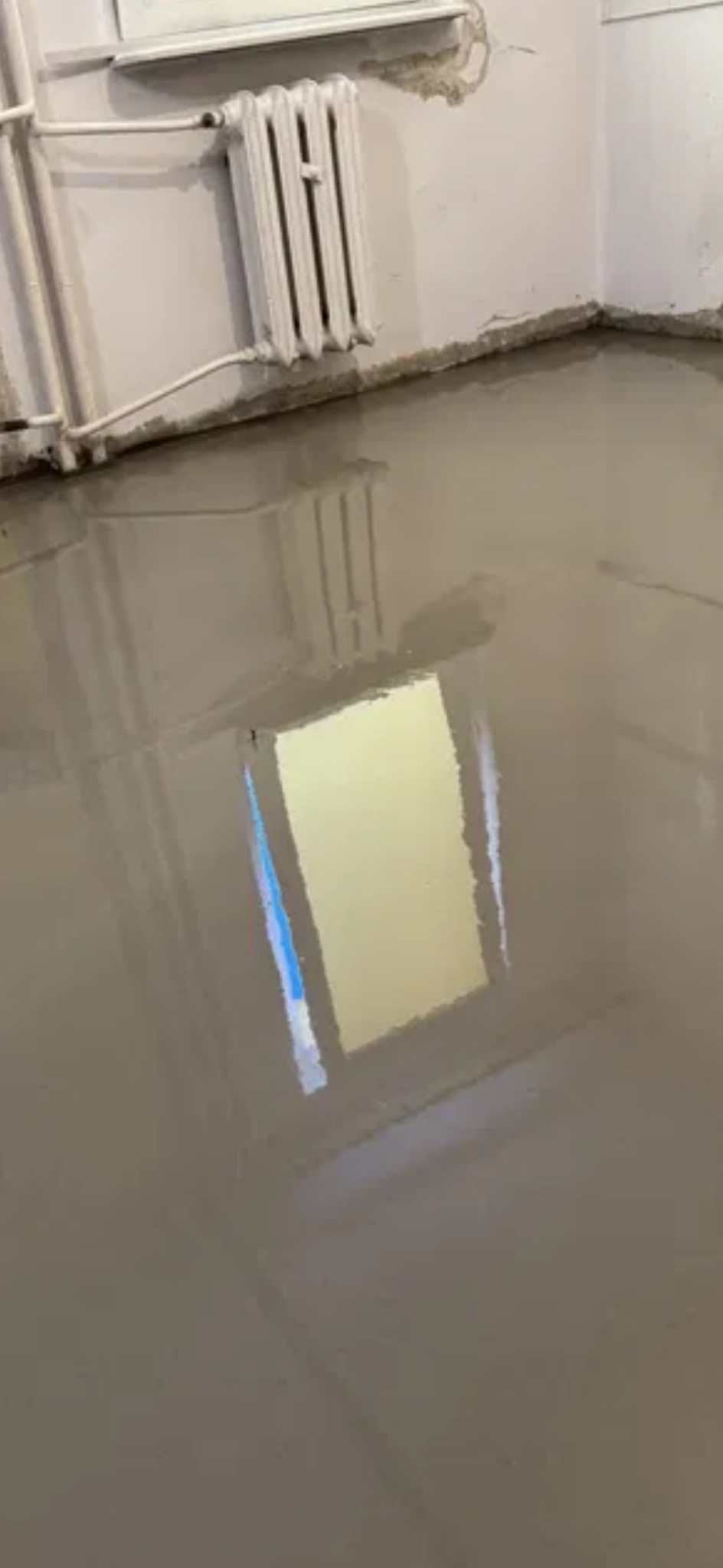Świeżo wylana, gładka posadzka żywiczna w pomieszczeniu z grzejnikiem żeberkowym. Widoczne ślady po zalaniu przy ścianach. Okno odbija się w mokrej powierzchni.