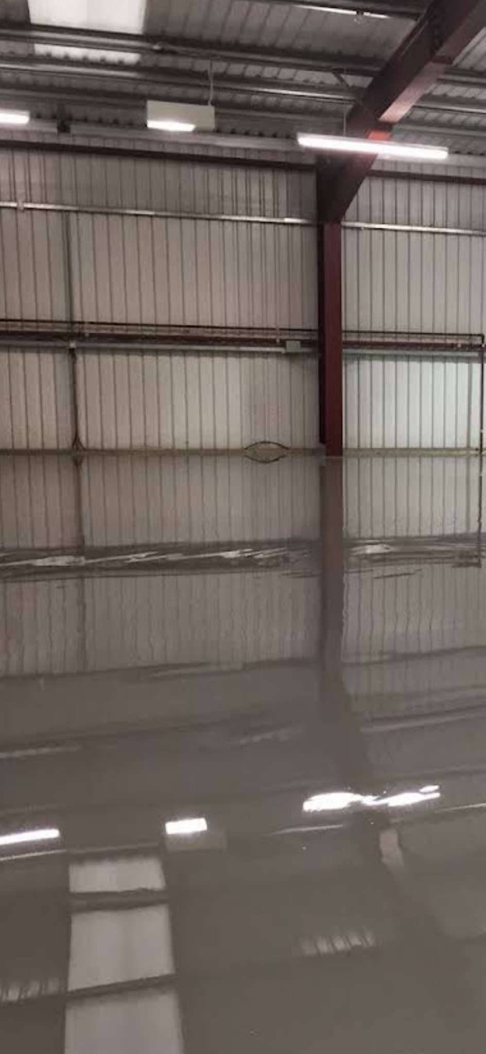 Błyszcząca, szara posadzka w hali przemysłowej z metalowymi ścianami i oświetleniem. Widoczne odbicia w gładkiej powierzchni podłogi.