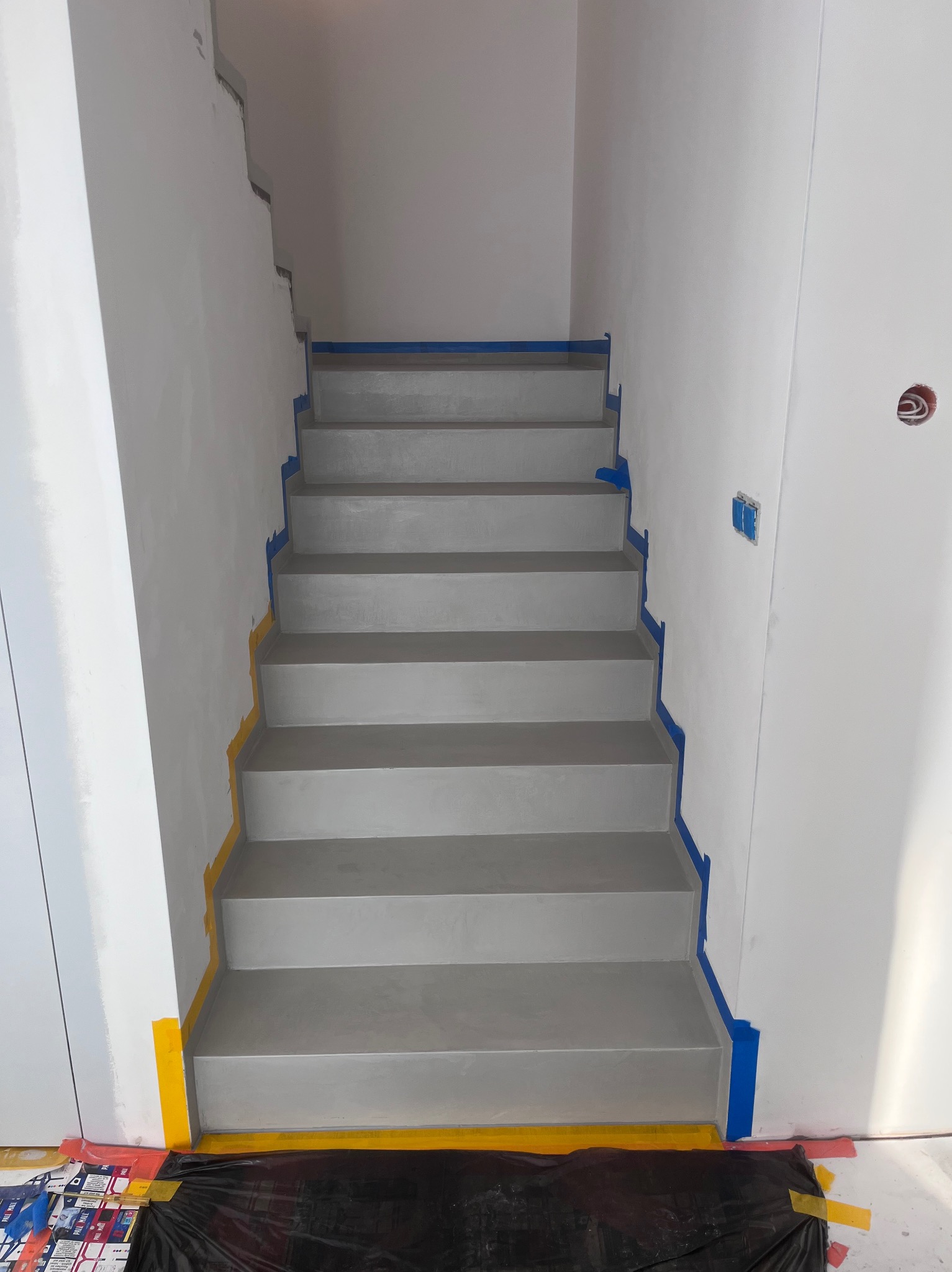 Schody wykończone szarą posadzką, zabezpieczone taśmą malarską. Widoczne detale wykończenia i przygotowania do dalszych prac. Minimalistyczny design.
